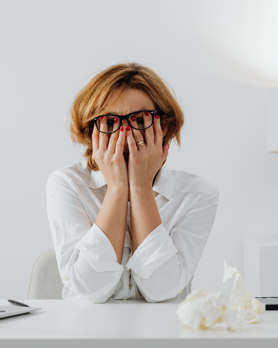 A woman with short red hair, wearing glasses and a white blouse, sitting at a desk with crumpled paper in front of her, holding her face in her hands and appearing stressed or overwhelmed.