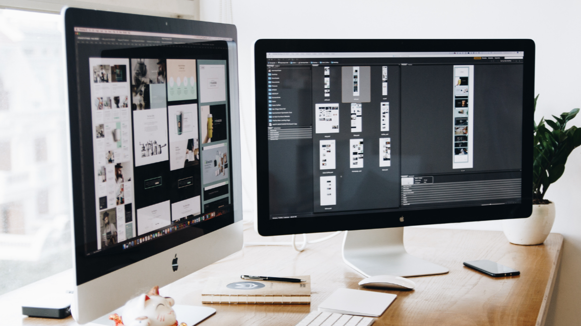 Two Apple iMac computers on a wooden desk, displaying design or photo editing projects, with a potted plant and a smartphone nearby.