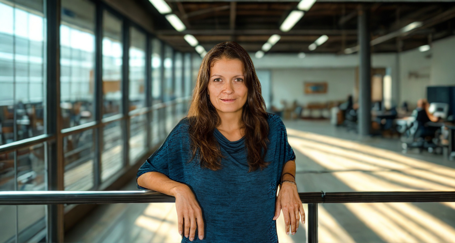 A woman with long brown hair wearing a blue shirt standing in a modern office building with glass walls and sunlight streaming in.