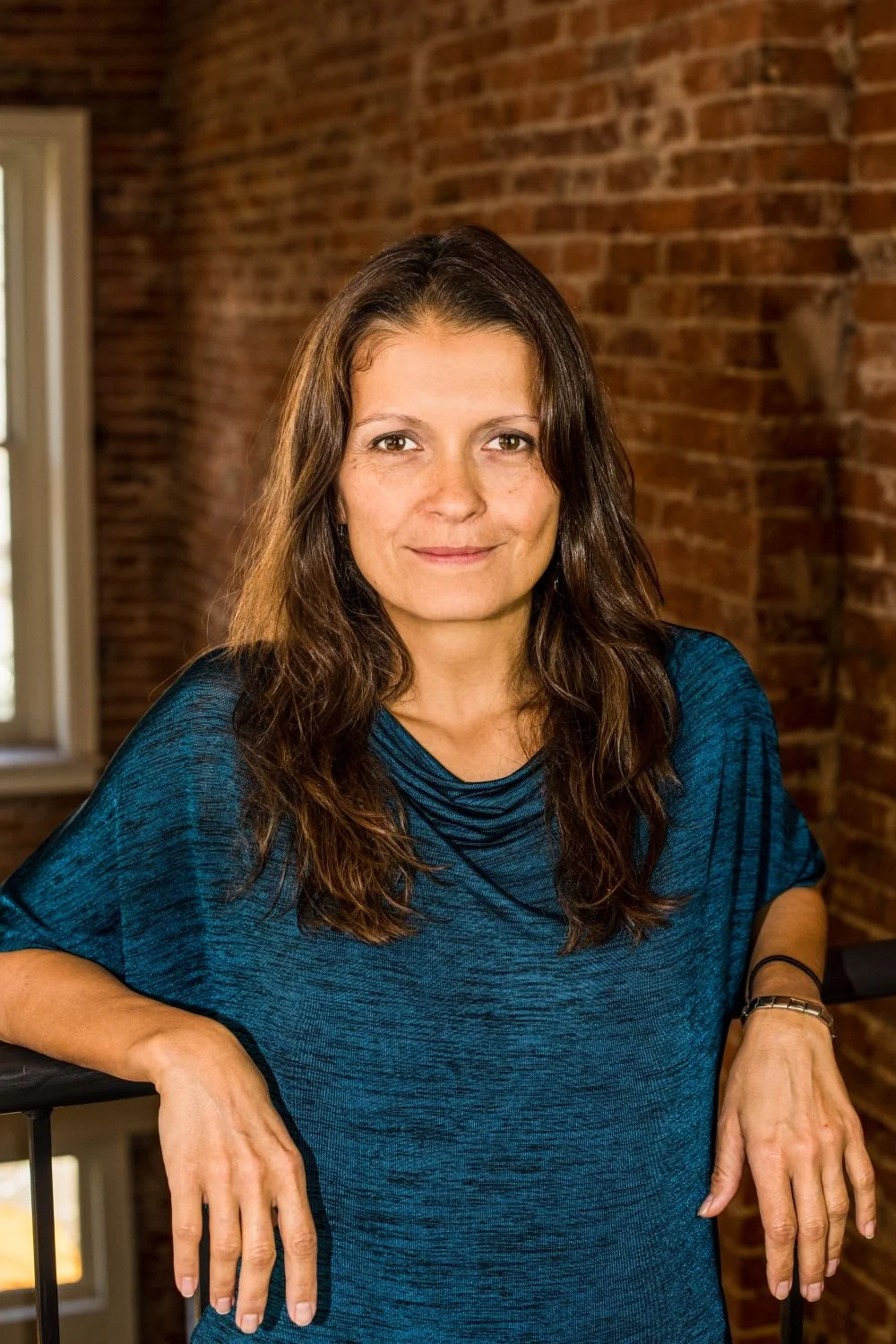 A woman, the owner of The BrandStory Connective, with long brown hair wearing a blue top, standing indoors with a brick wall in the background.