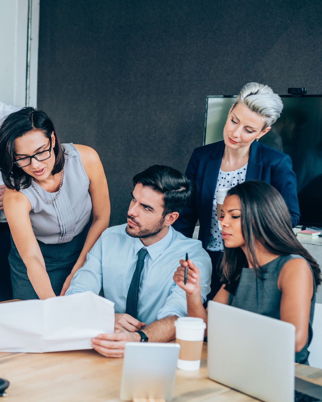 Five professionals in a business meeting around a table, discussing a document, with laptops and coffee cups present.