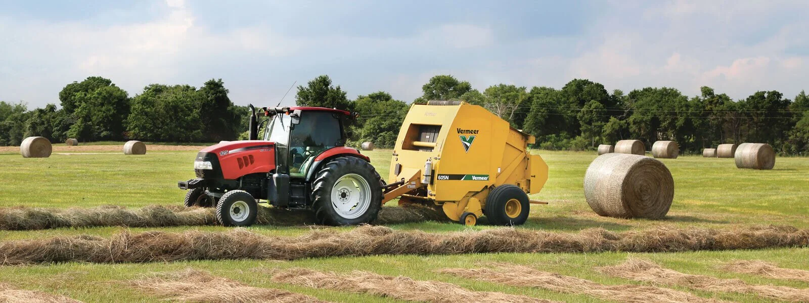 A tractor pulling a hay baler in a field with large round hay bales and trees in the background.