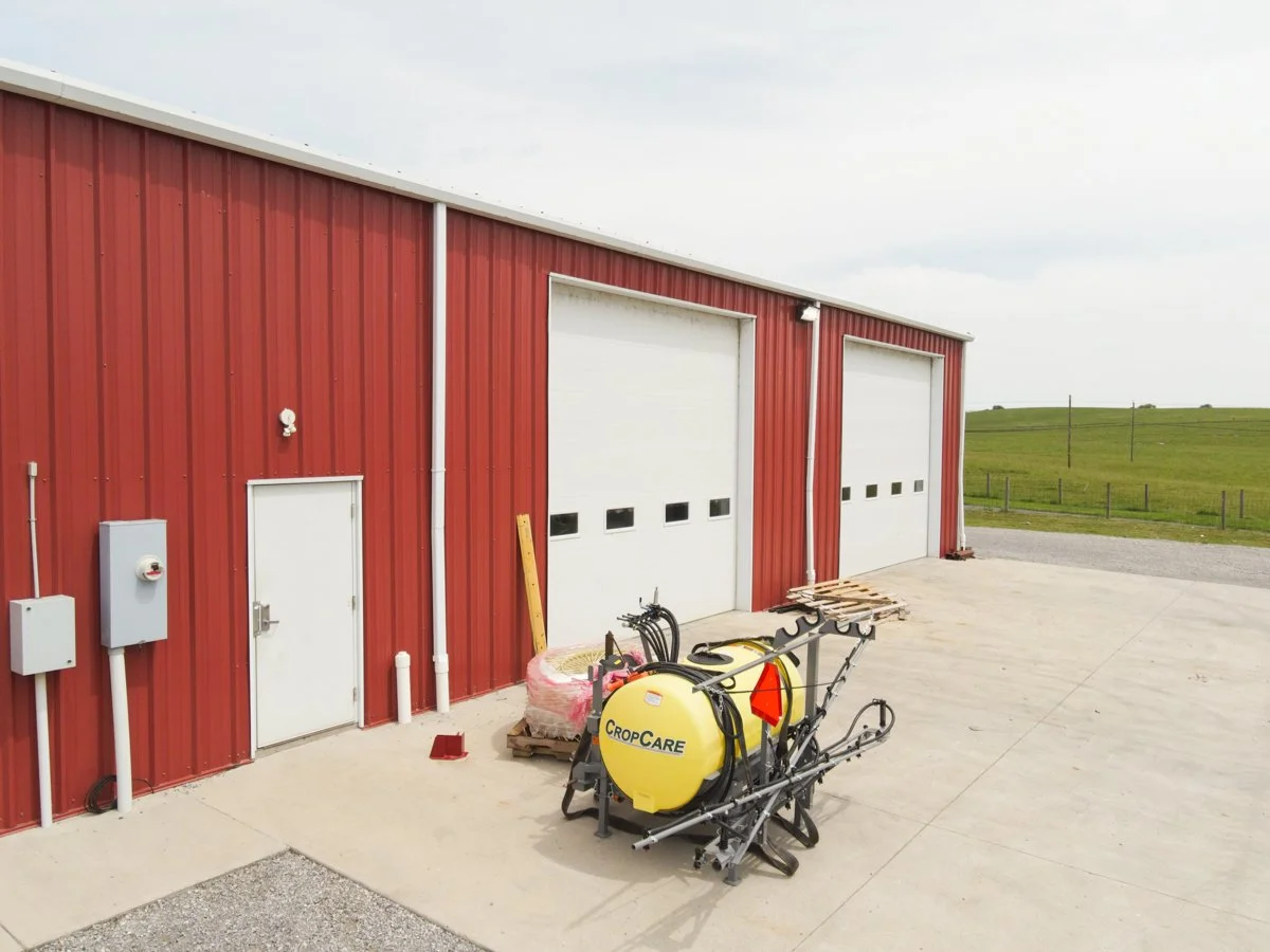 Exterior of a red farm building with two large white garage doors, a smaller white door, and farm equipment including a yellow Sprayer labeled 'Crop Care'.