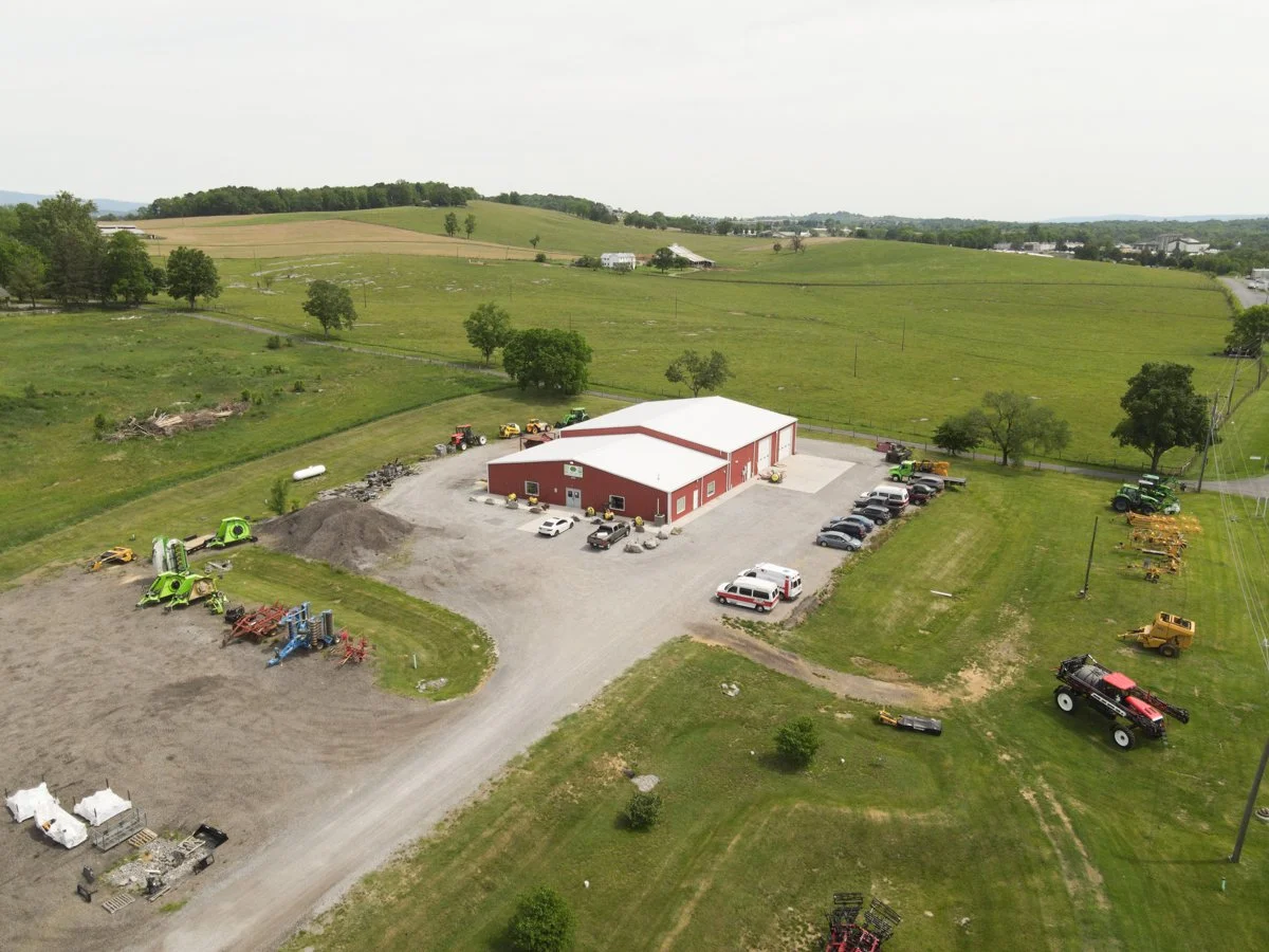 A rural farm scene with a red barn, various farm equipment, tractors, and vehicles parked near a gravel driveway and surrounding green fields and rolling hills.