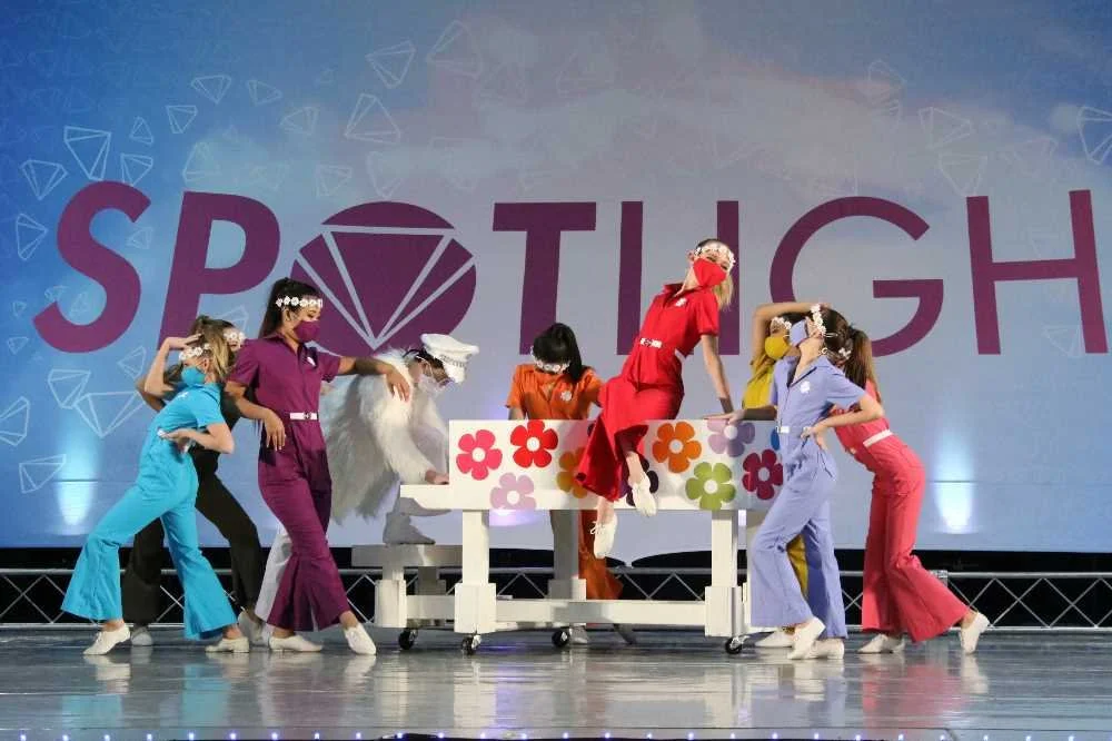 Children dressed in colorful scrubs and costumes performing on stage with a 'SPOTLIGHT' backdrop, engaging with a prop table with floral decorations.