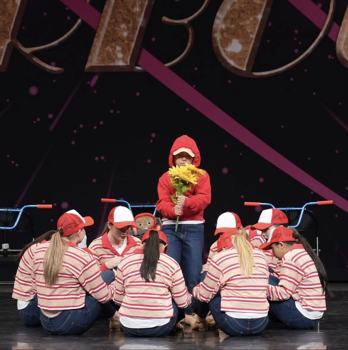 A group of children wearing identical red and white striped shirts, red caps, and jeans are kneeling in a circle on stage. One child stands in the middle holding a bouquet of yellow flowers, wearing a red hoodie and a baseball cap. Behind them, a per