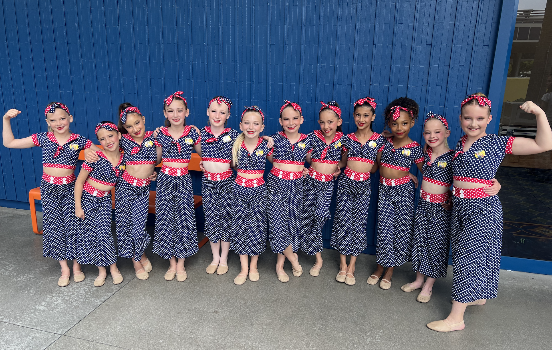 A group of young girls dressed in matching red, white, and blue polka dot costumes, standing arm in arm in front of a blue wall, with some posing with biceps flexed and smiling.