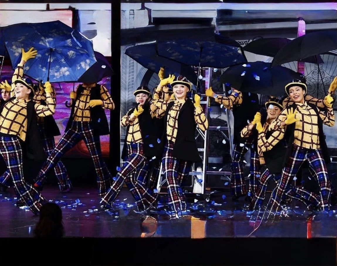 Group of female performers dressed in yellow plaid tops, plaid pants, and black vests, holding black umbrellas with a blue sky pattern, dancing on stage with confetti on the floor.