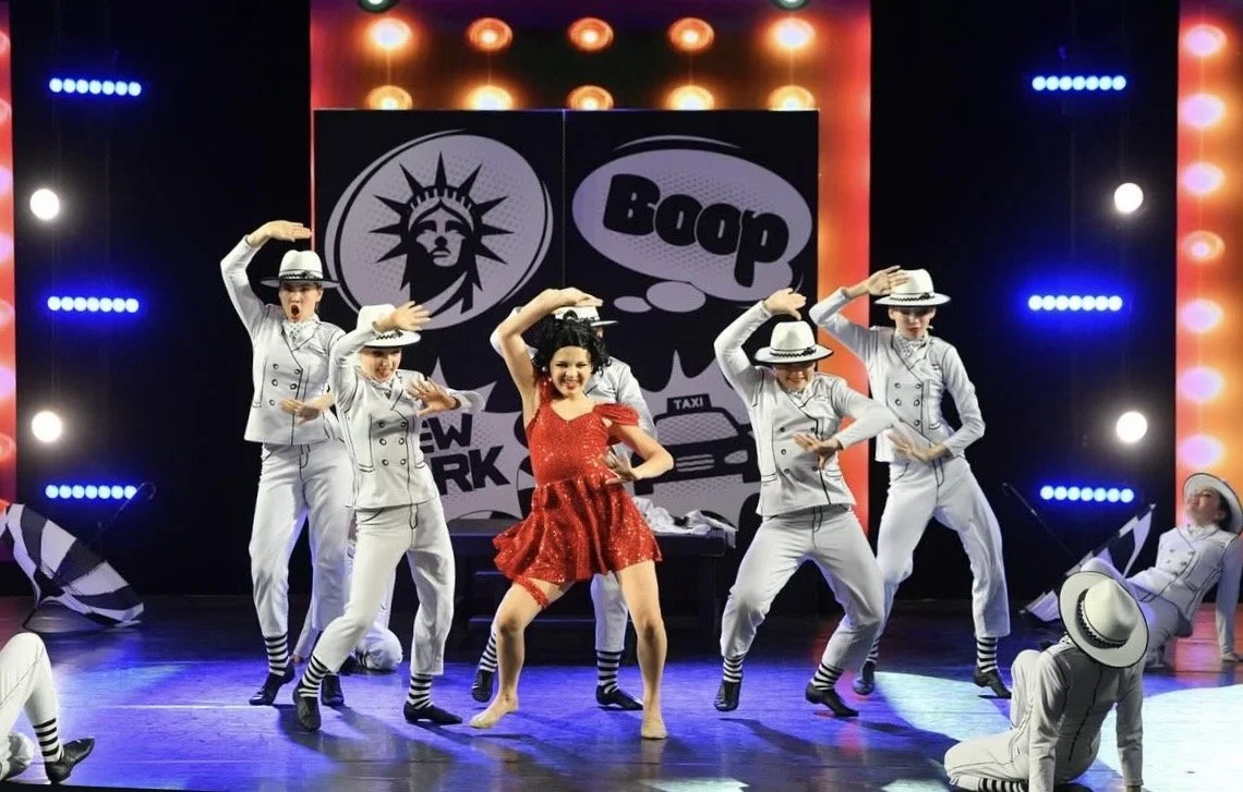 Group of dancers performing on stage, with a woman in a red dress in the center and men in gray suits and white hats around her, stage decorated with bright lights and comic-style graphics in the background.