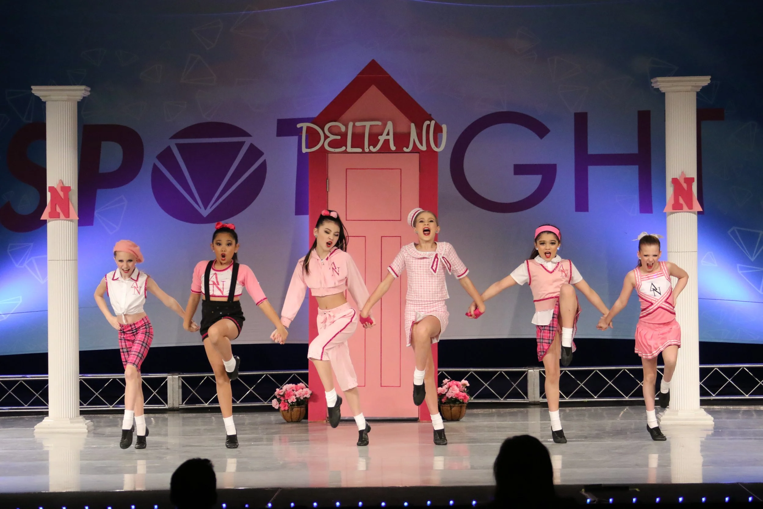 Group of young girls in pink and white costumes performing a dance on stage, with a backdrop that has the words 'SPO' and 'DELTA NU' and decorative elements resembling a school setting.