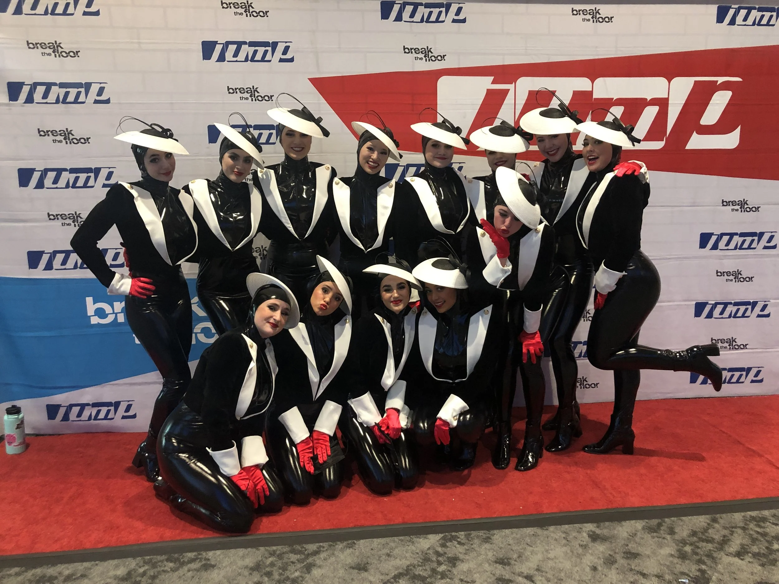 Group of women dressed as stewards with black, white, and red uniforms and large white hats, posing in front of a backdrop with logos for 'Break the Floor' and 'Jump'.