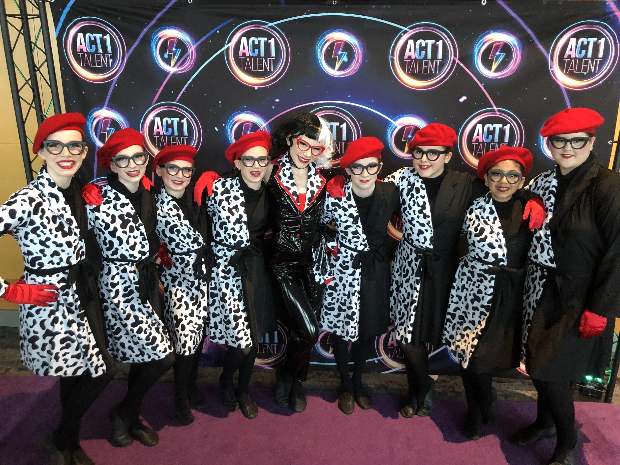 A group of nine performers dressed in Dalmatian print coats, red berets, black glasses, and red gloves standing together in front of a backdrop that reads 'ACT1 TALENT' with neon-style logos and lightning bolt symbols.