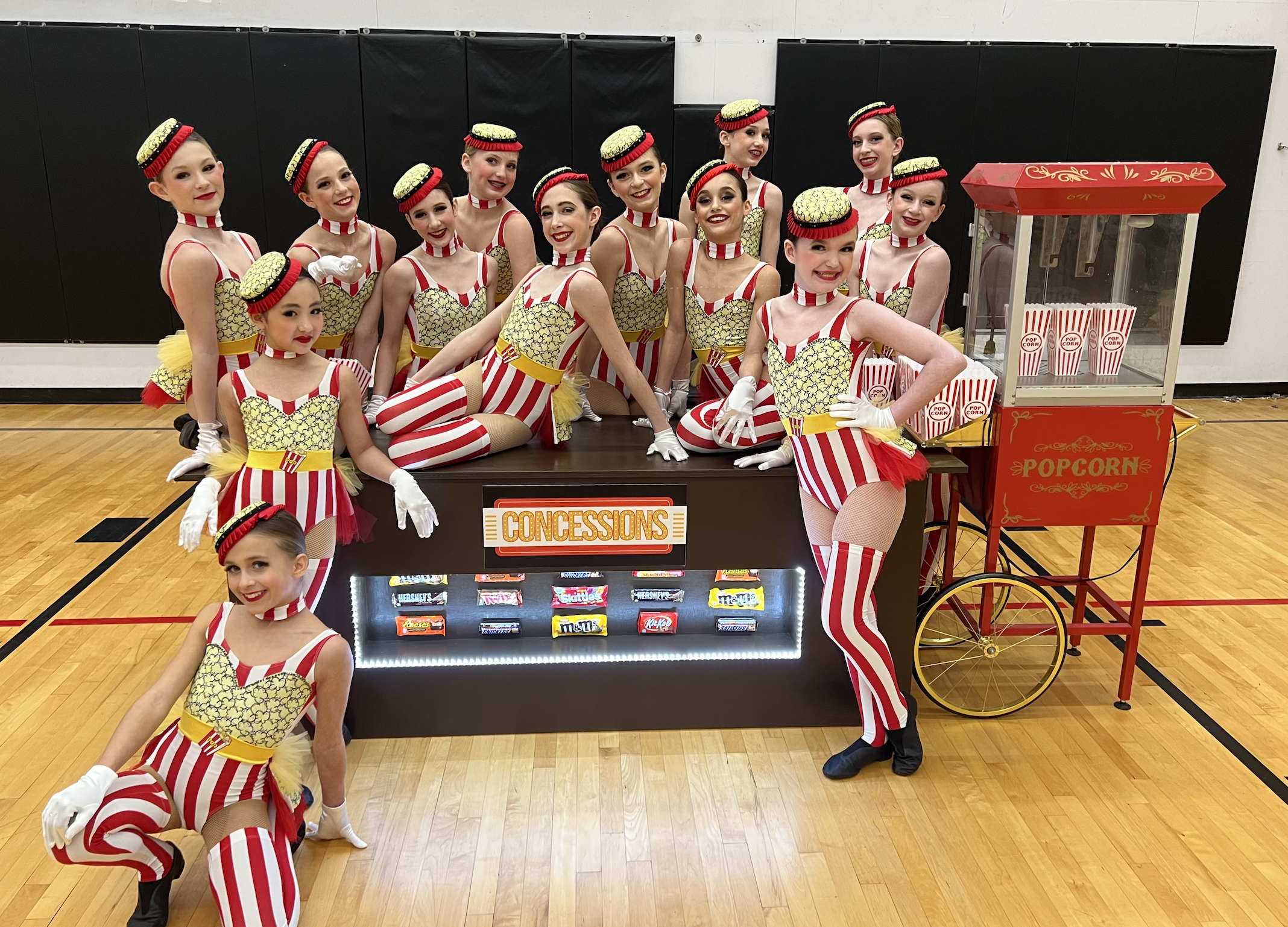 A group of young female performers dressed as popcorn with red and white striped costumes, yellow accents, and popcorn hat headpieces, posing next to a popcorn vending cart in a gymnasium.