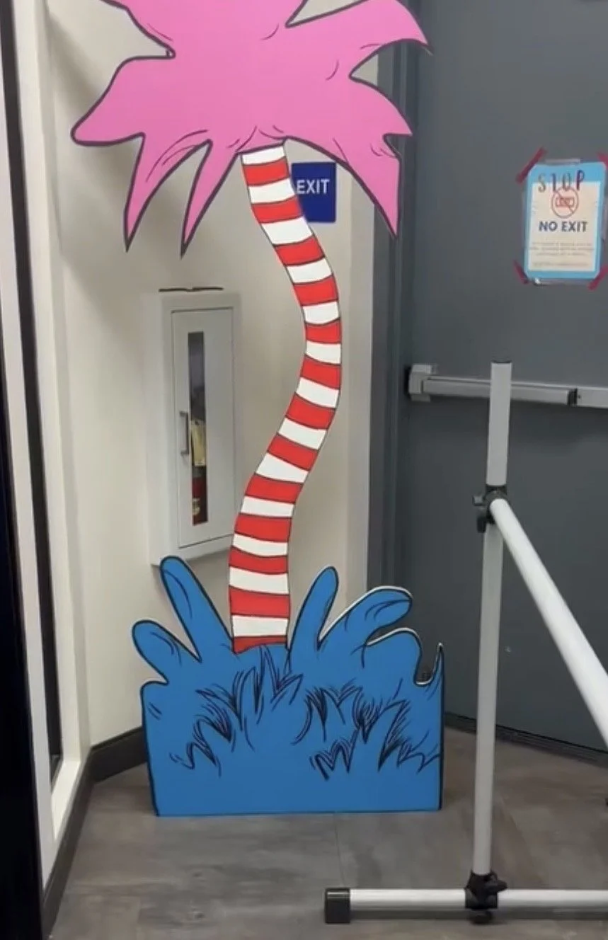 Cartoon cutout of a colorful plant with pink spiky leaves and a striped red and white stem, placed in a blue base with grass-like design, next to a fire extinguisher cabinet and exit signs.