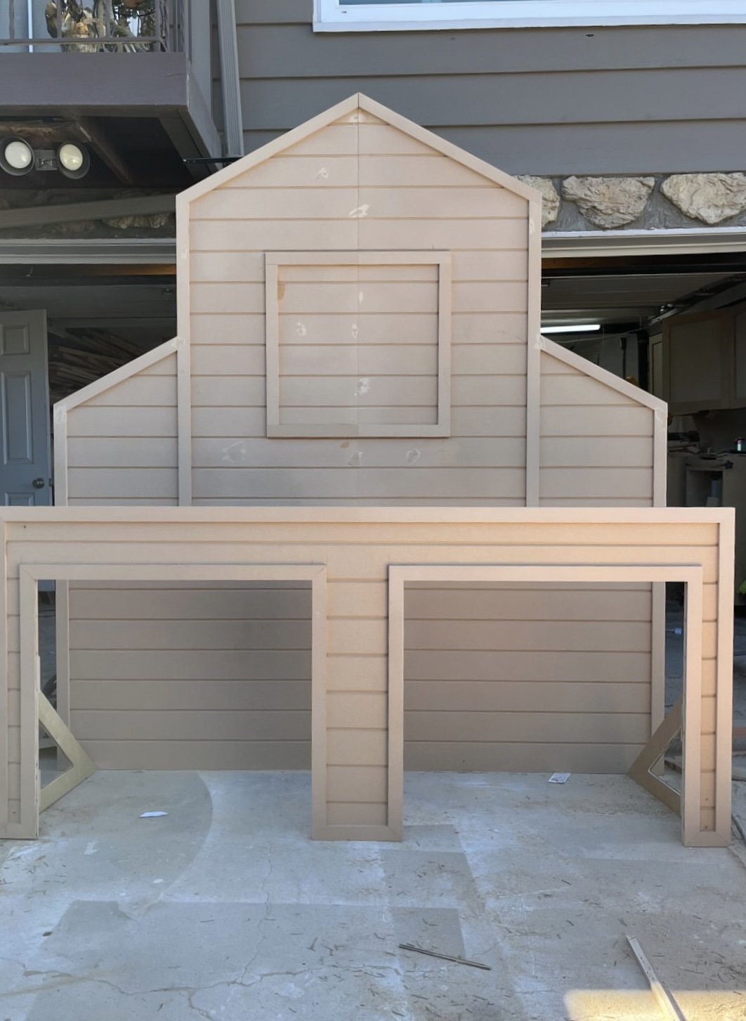 Unfinished wooden playhouse frame with horizontal siding and a central window frame, sitting outdoors on a concrete surface.