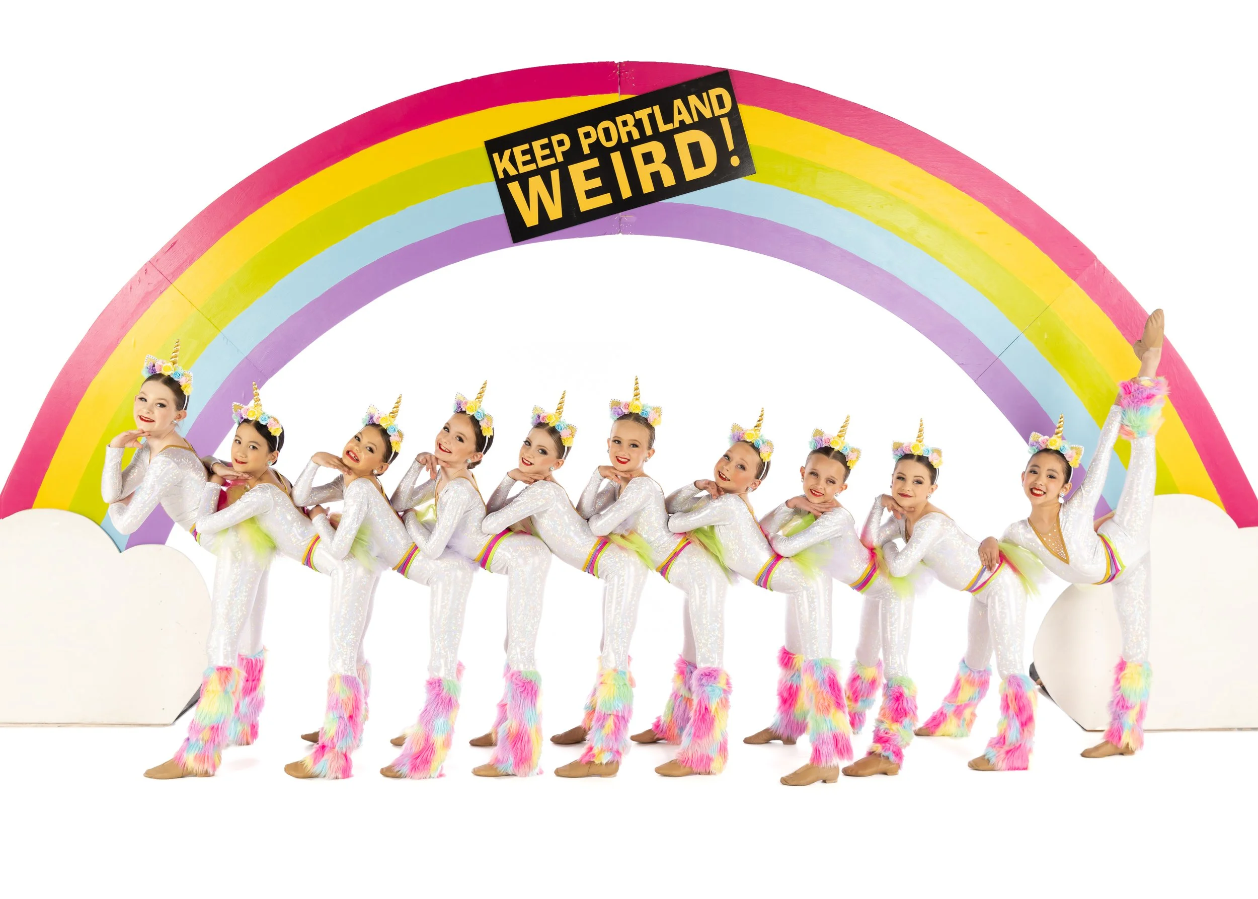 Line of young girls in unicorn costumes with rainbow-colored fur leg warmers, standing under a rainbow display with a sign that says 'Keep Portland Weird!'