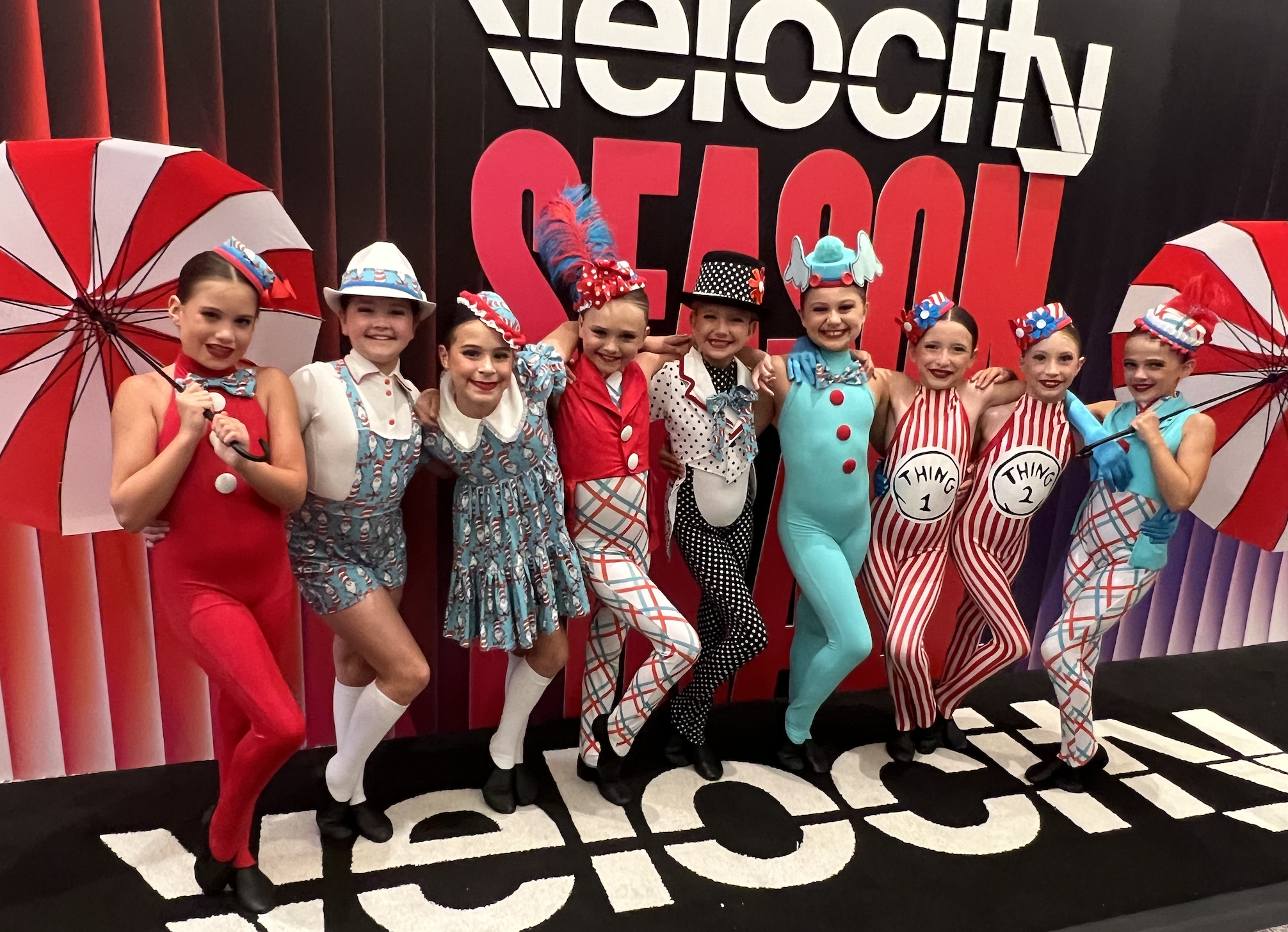 A group of children dressed in colorful clown costumes, standing together and smiling, against a background with large red and white letters and umbrellas.