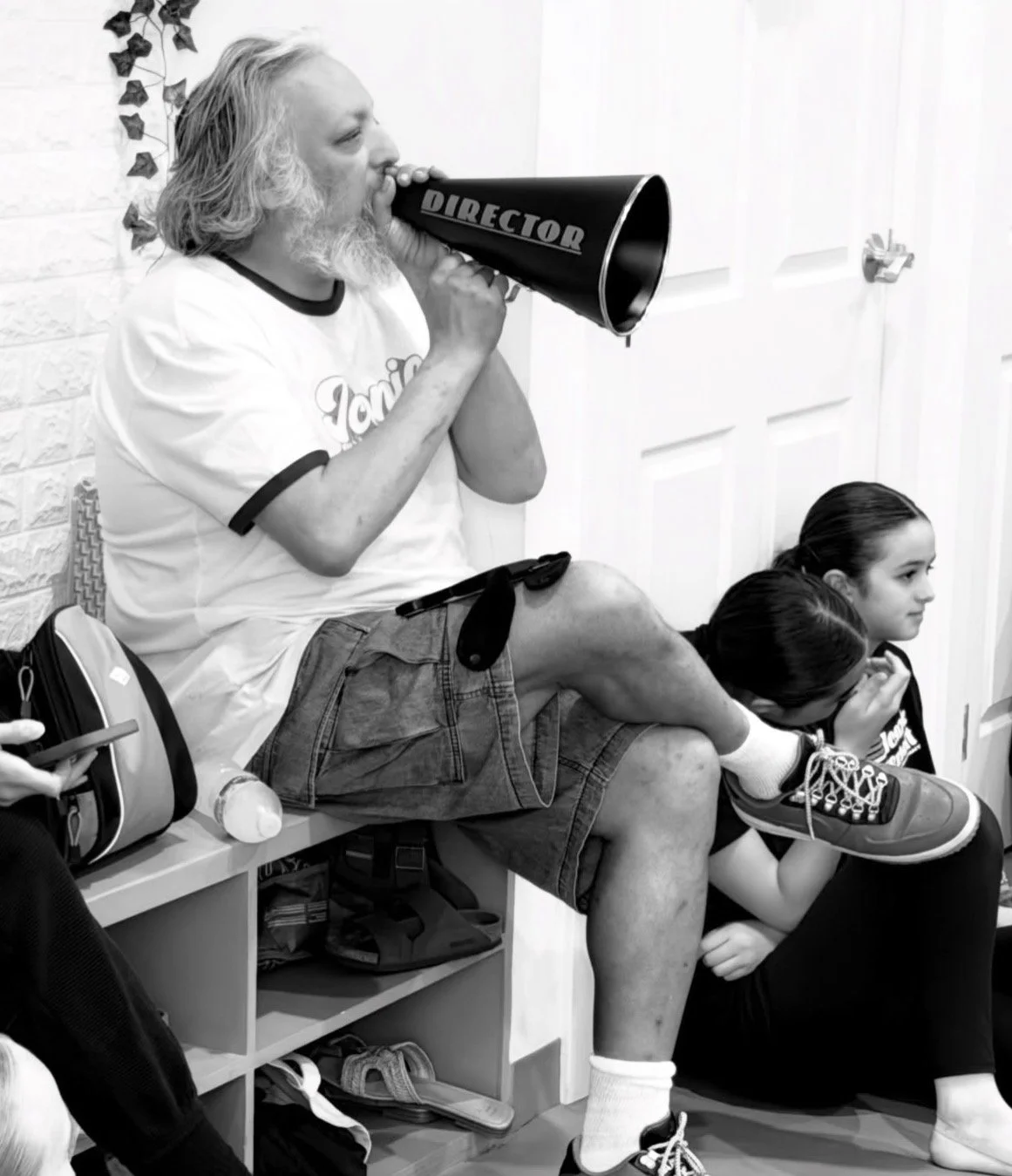 A man with long gray hair and a beard sitting on a bench, holding a megaphone labeled 'DIRECTOR' to his mouth. He's wearing a t-shirt and shorts. Two young girls sit beside him, one with her head down and the other looking ahead, both in casual clothing, in what appears to be a waiting area.