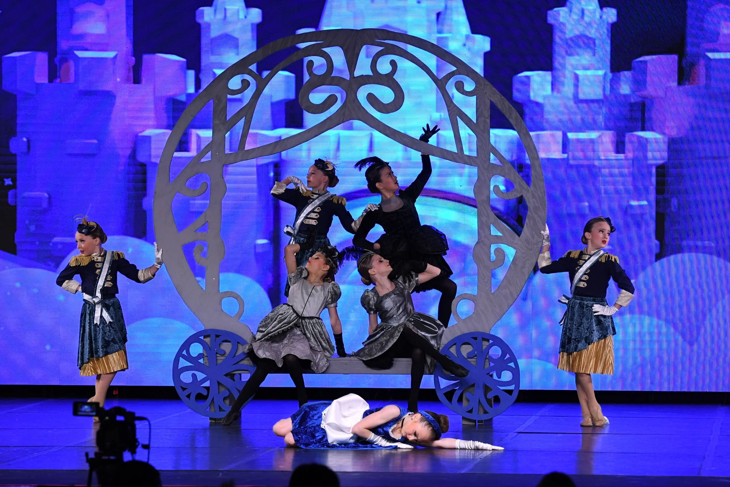 Children in historical costumes perform on stage with a large decorative carriage and castle backdrop, engaging in a theatrical or dance performance.