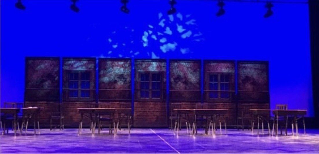 Empty stage with wooden tables and chairs, blue lighting, and a painted backdrop of trees and house windows