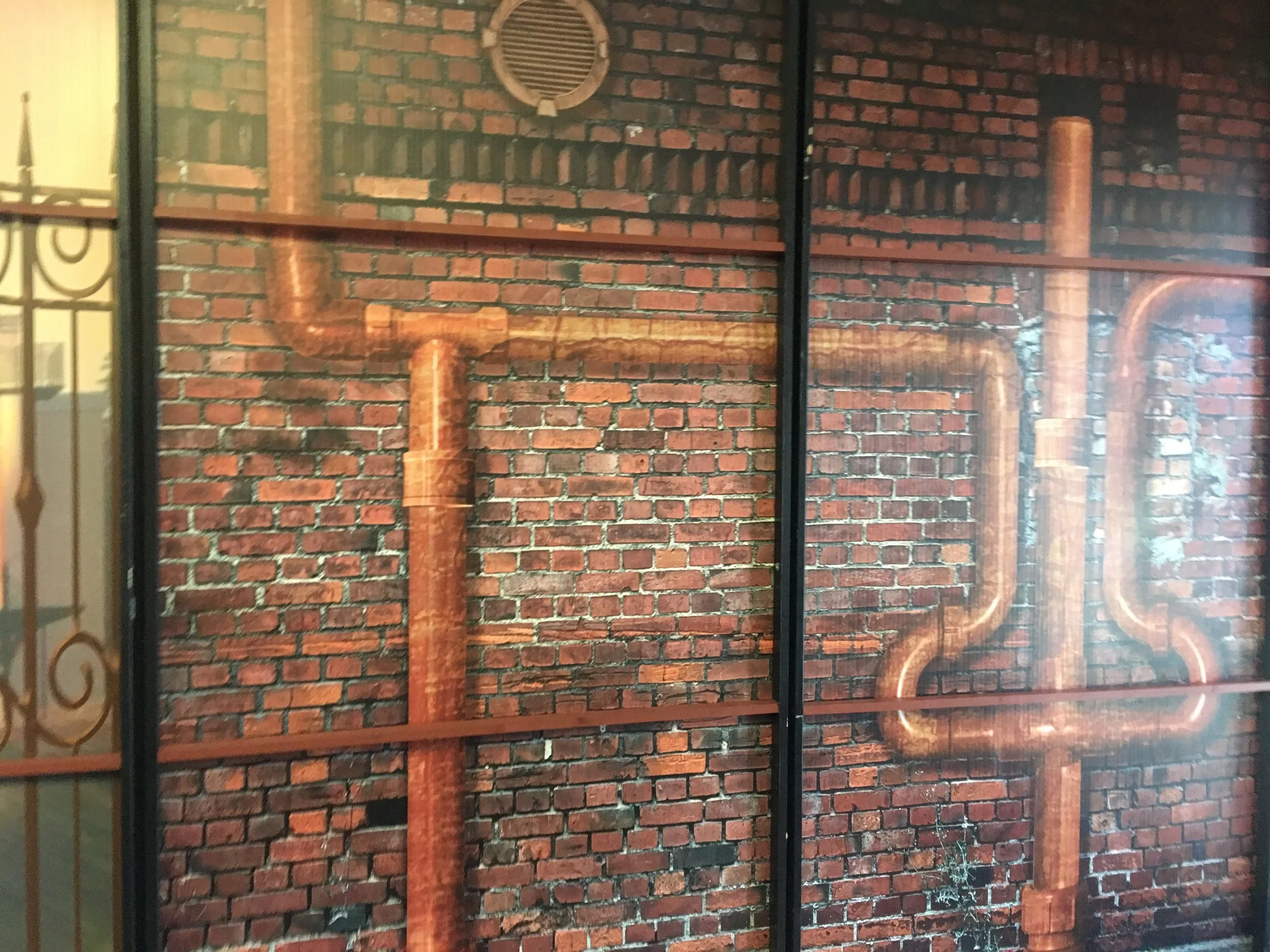 A brick wall with copper pipes and a round vent, viewed through a glass window with a black border.