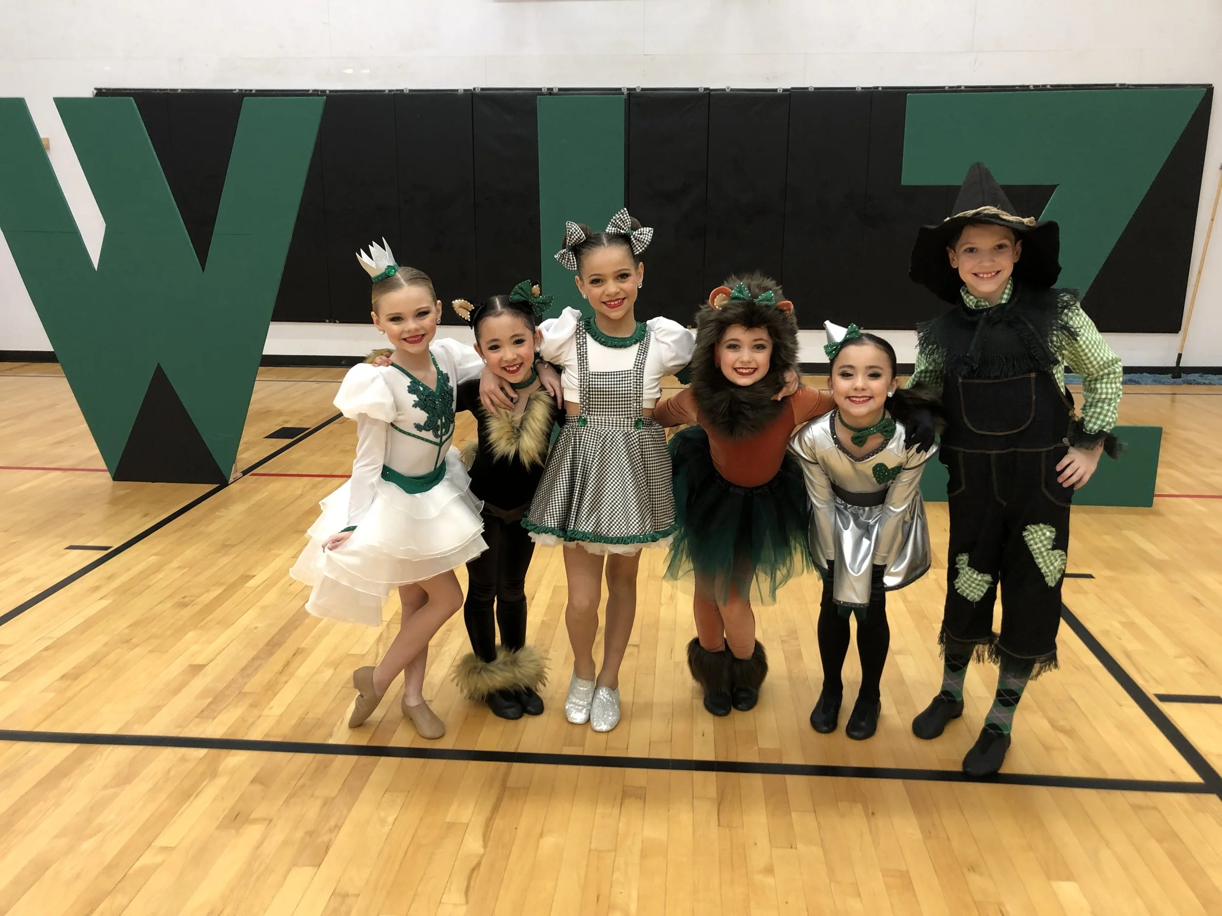 A group of six children dressed in costumes, posing in a gymnasium with a black and green wall and large green and black letters in the background.