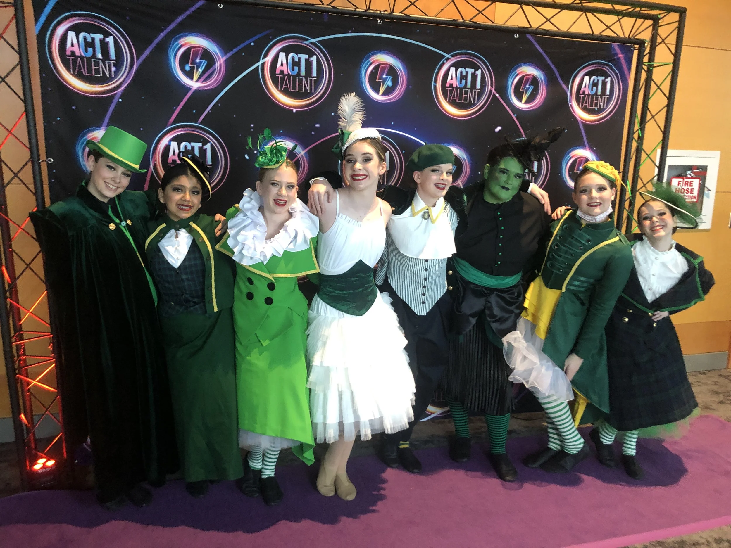 Group of seven performers wearing colorful costumes, some with hats and accessories, standing in front of a backdrop with 'ACT 1 TALENT' logos.
