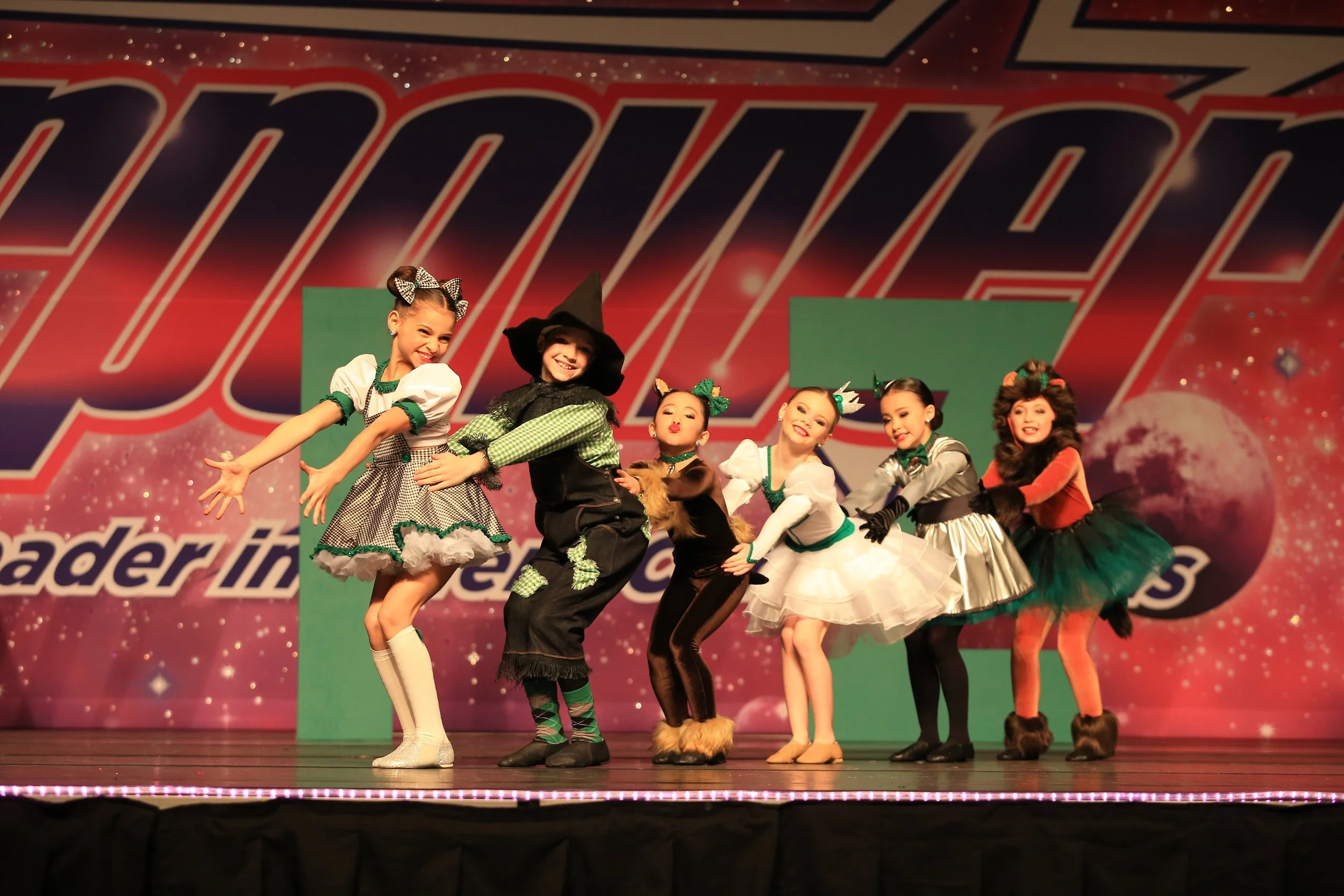 Children dressed in costumes performing a dance on stage with a pink and purple background and the words "Leader In" visible.