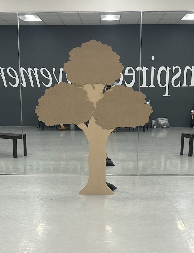 A cardboard cutout of a tree is positioned in front of a mirrored wall, reflecting the room behind it.