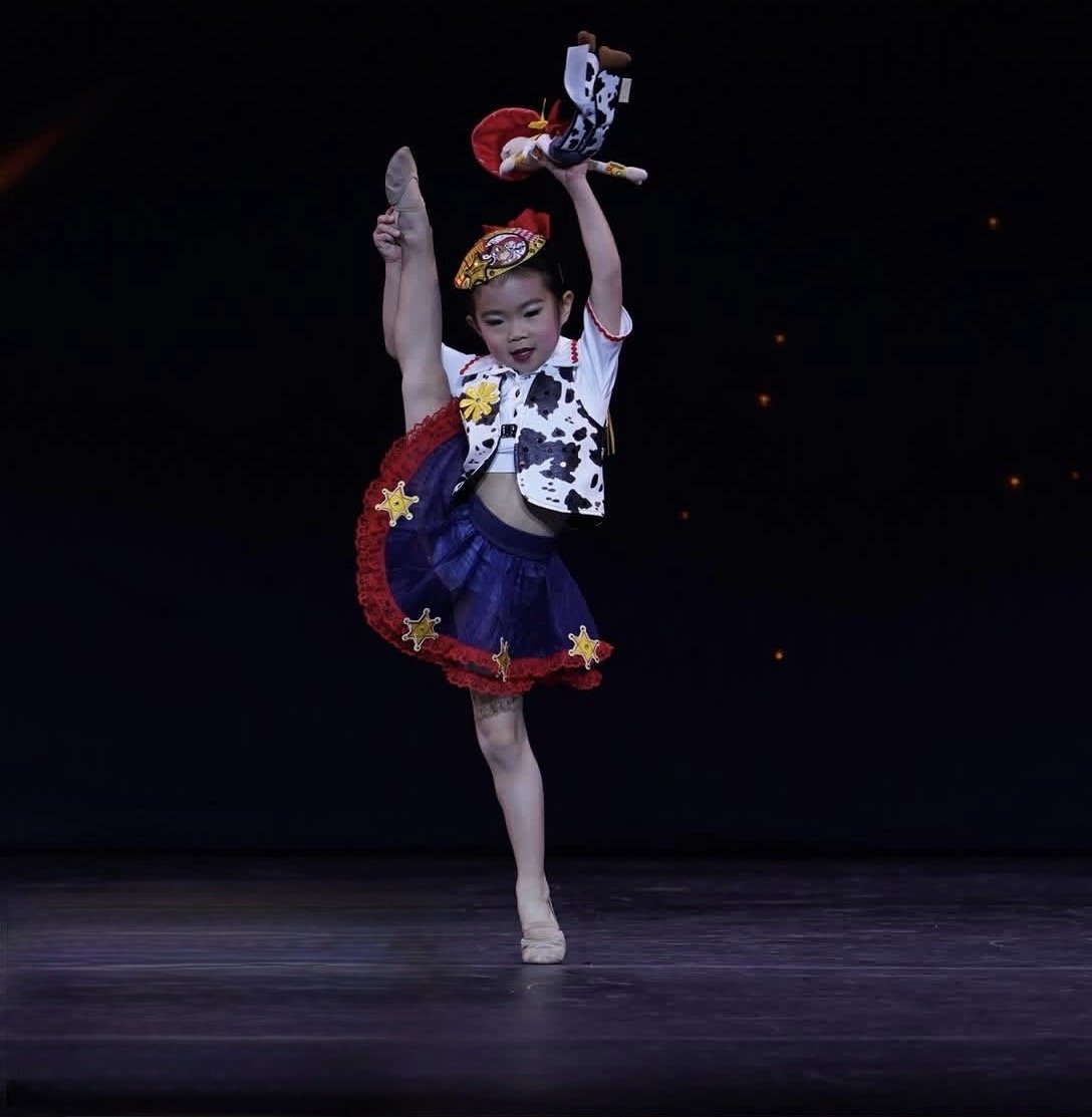 Young girl dancing in a colorful costume on stage against a dark background.