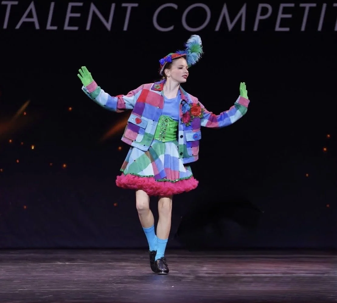 A performer on stage in colorful, eclectic costume with a plaid jacket, pink fur trim skirt, green corset, and a feathered hat, striking a pose with arms outstretched. The stage background has partial text reading 'TALENT COMPET'.