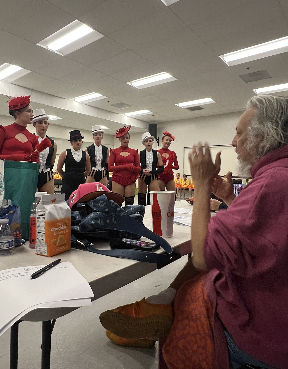 Group of performers in colorful costumes standing in line for a rehearsal or show, with a man sitting at a table observing them.