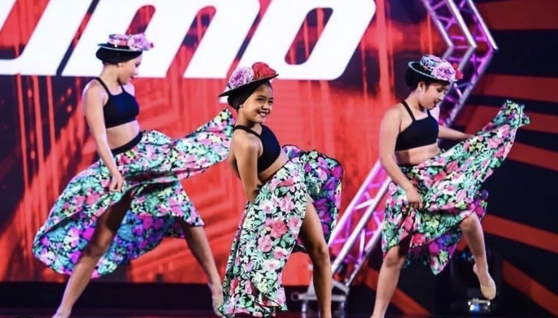 Three young girls in black crop tops and floral skirts performing a dance on stage, with pink and purple hats. They are smiling and holding their skirts as part of their dance routine.