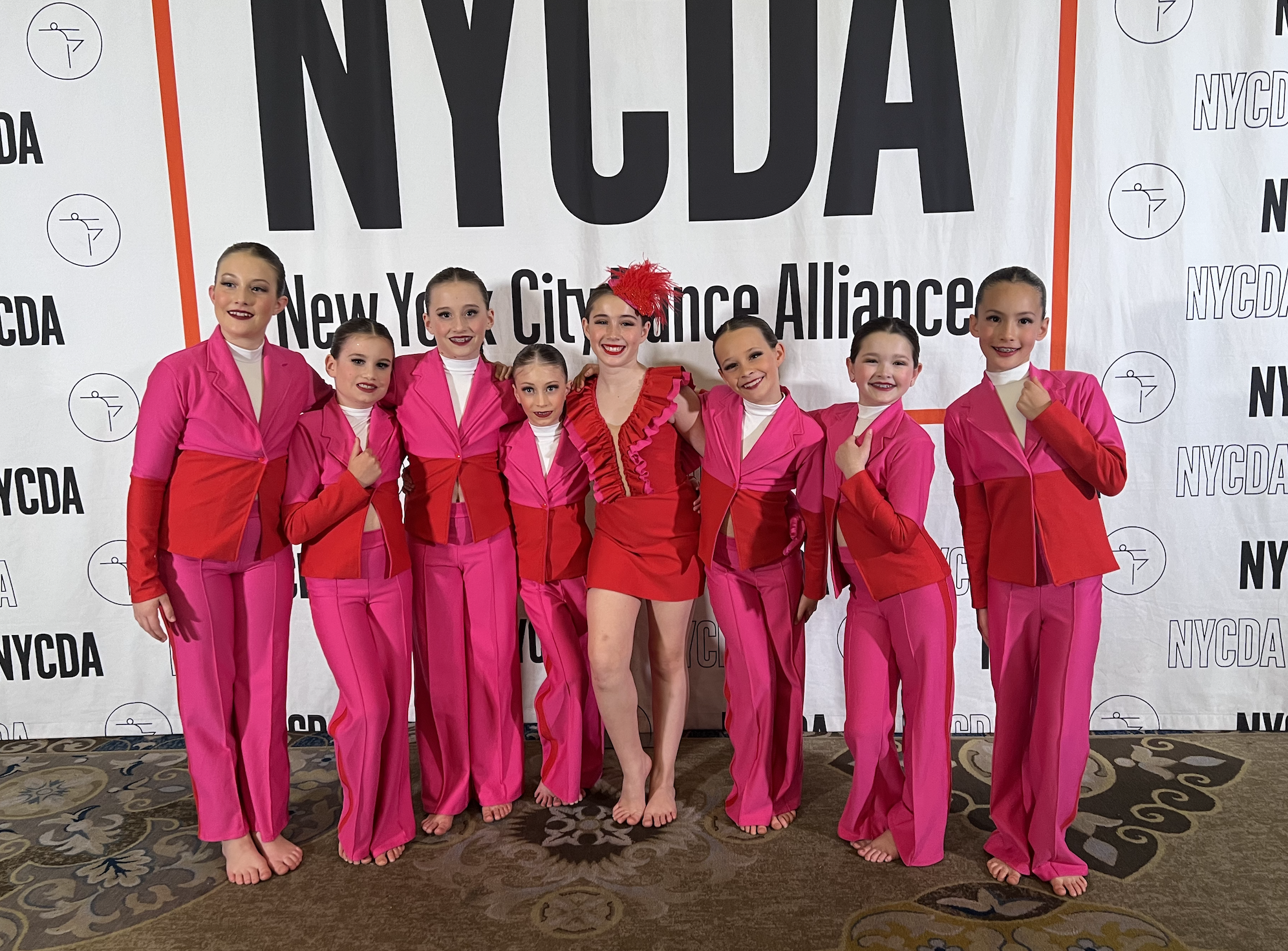 Group of young girls in pink costumes standing together in front of a NYCDTA banner at a dance event, smiling.