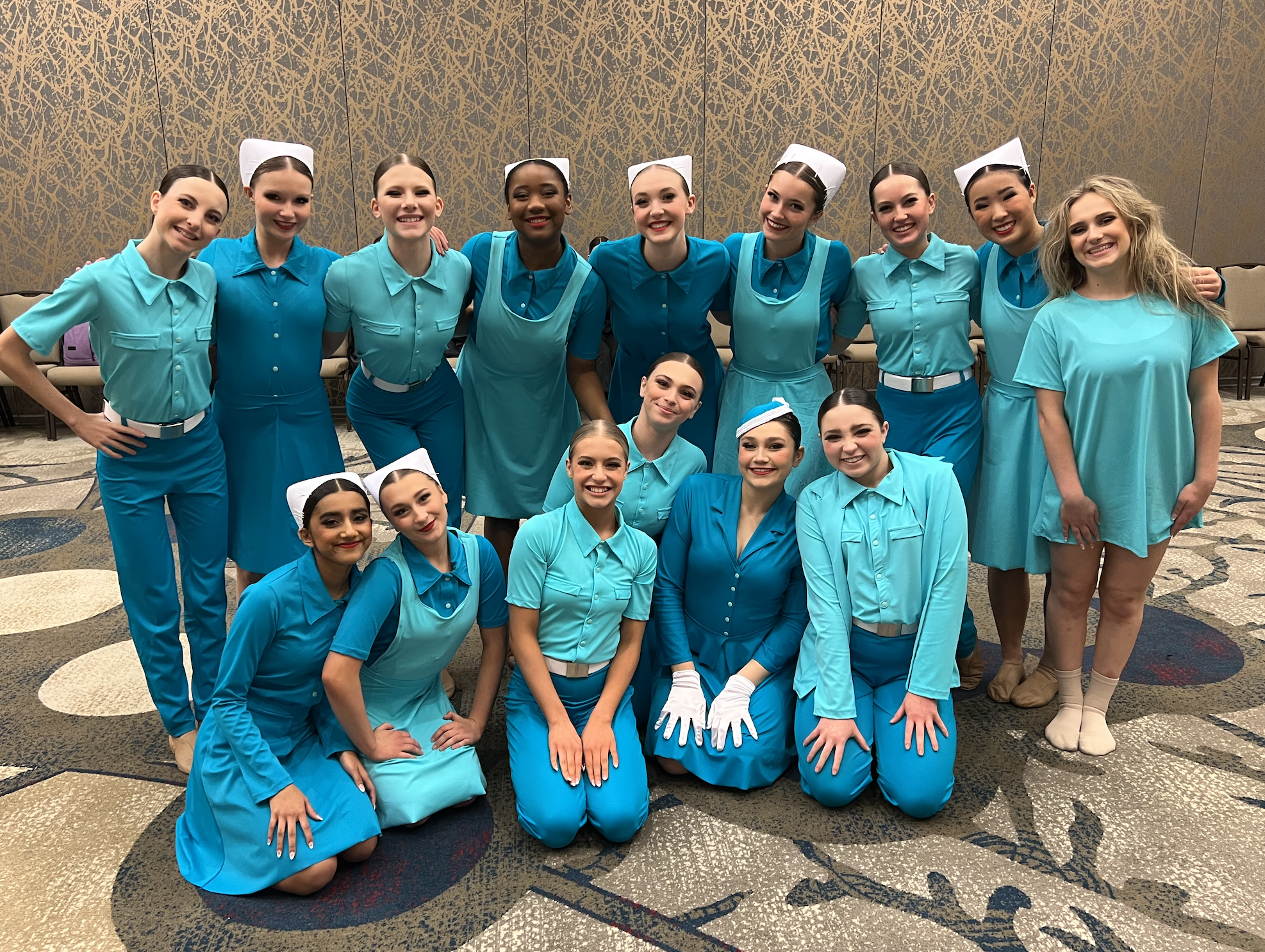 A group of young female dancers in blue costumes with white gloves and nurses' caps, posing together in a ballroom with a patterned wall.