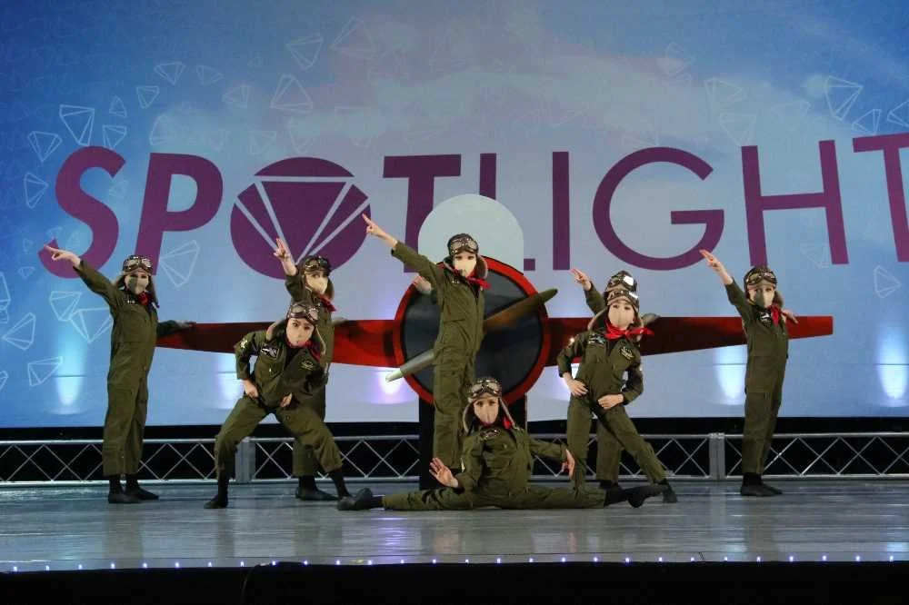Group of children dressed as pilots performing a dance or skit on stage with a backdrop that says 'SPOTLIGHT' with a diamond emblem.