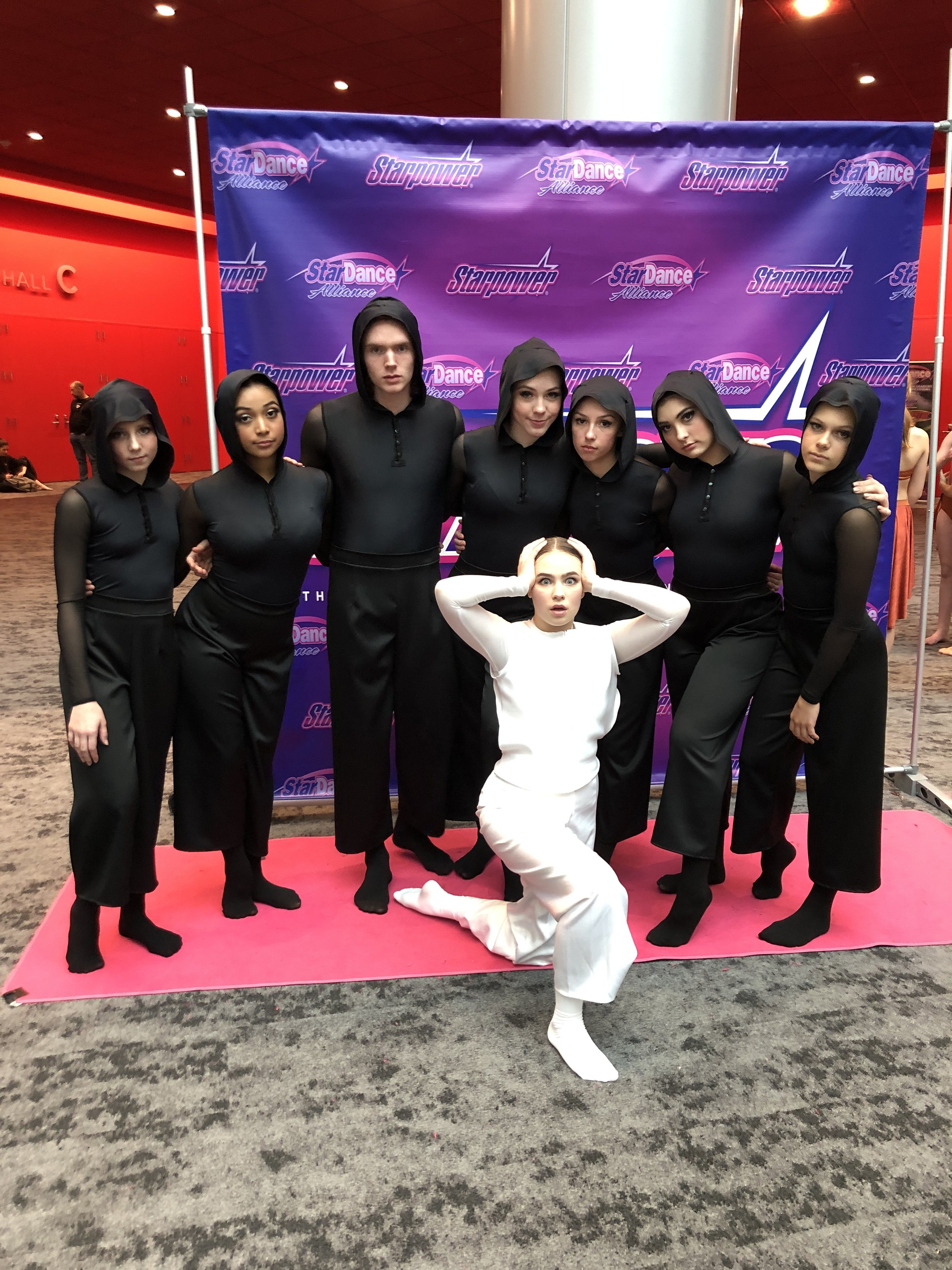 Group of six young women in black outfits with hoods on a pink carpet in front of a purple backdrop with 'StarDance Alliance' logos, with one woman kneeling in front wearing white.