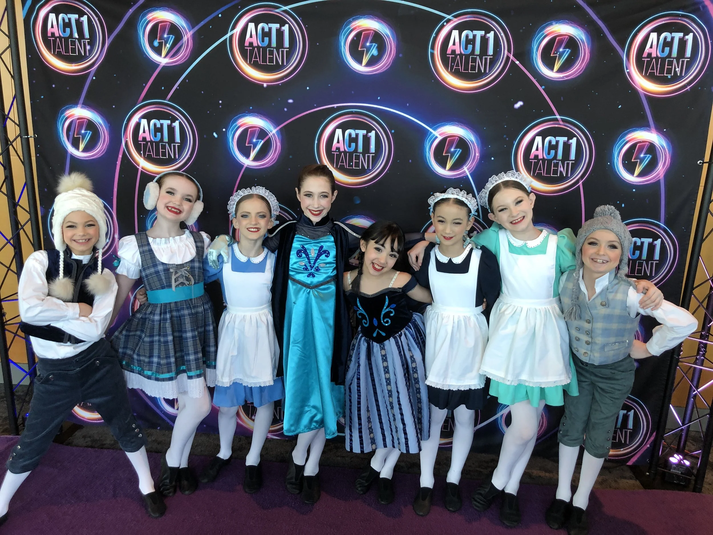 Group of young girls in colorful costumes standing in front of a backdrop with 'ACT 1 Talent' logos.