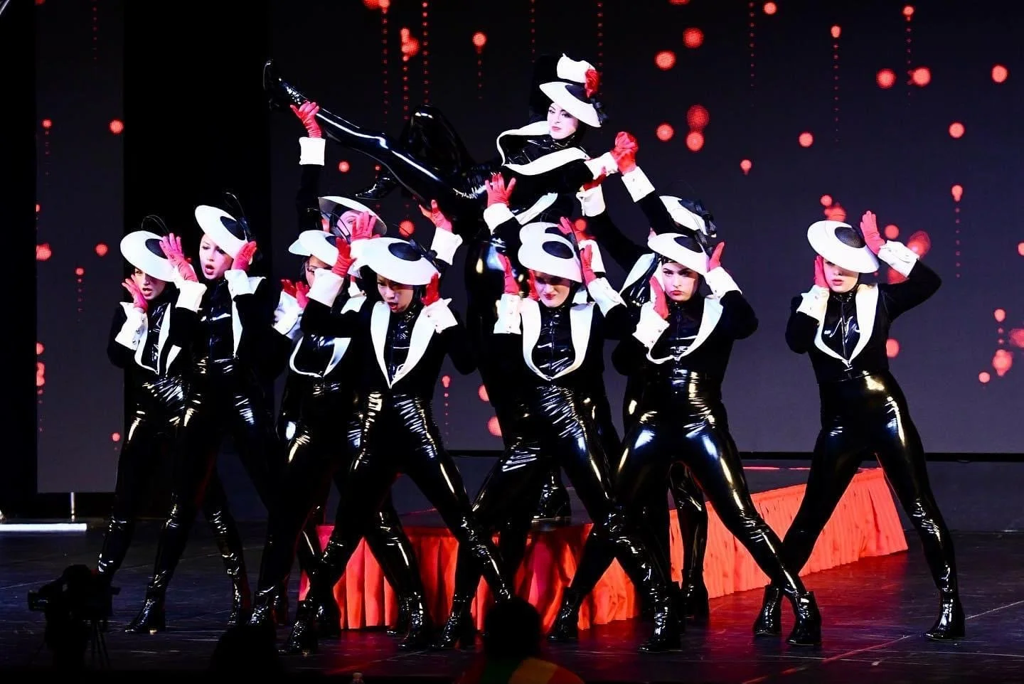 A dance performance on stage featuring nine women dressed in black shiny outfits with white accents, wearing large white hats with black spots, and pink gloves. One woman is lifted above the others in a pose, with their background displaying a digita