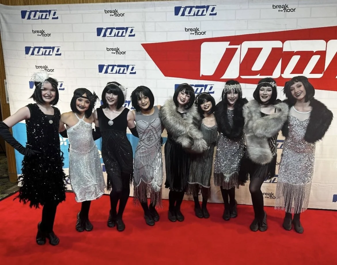 A group of nine women dressed in vintage flapper-style dresses and accessories, posing together on a red carpet in front of a backdrop with logos that say 'VOMP' and 'break the floor'.