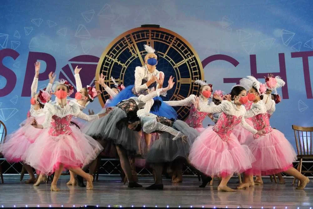 Ballet performers on stage wearing pink and gray tutus, with masks, in front of a large clock background.