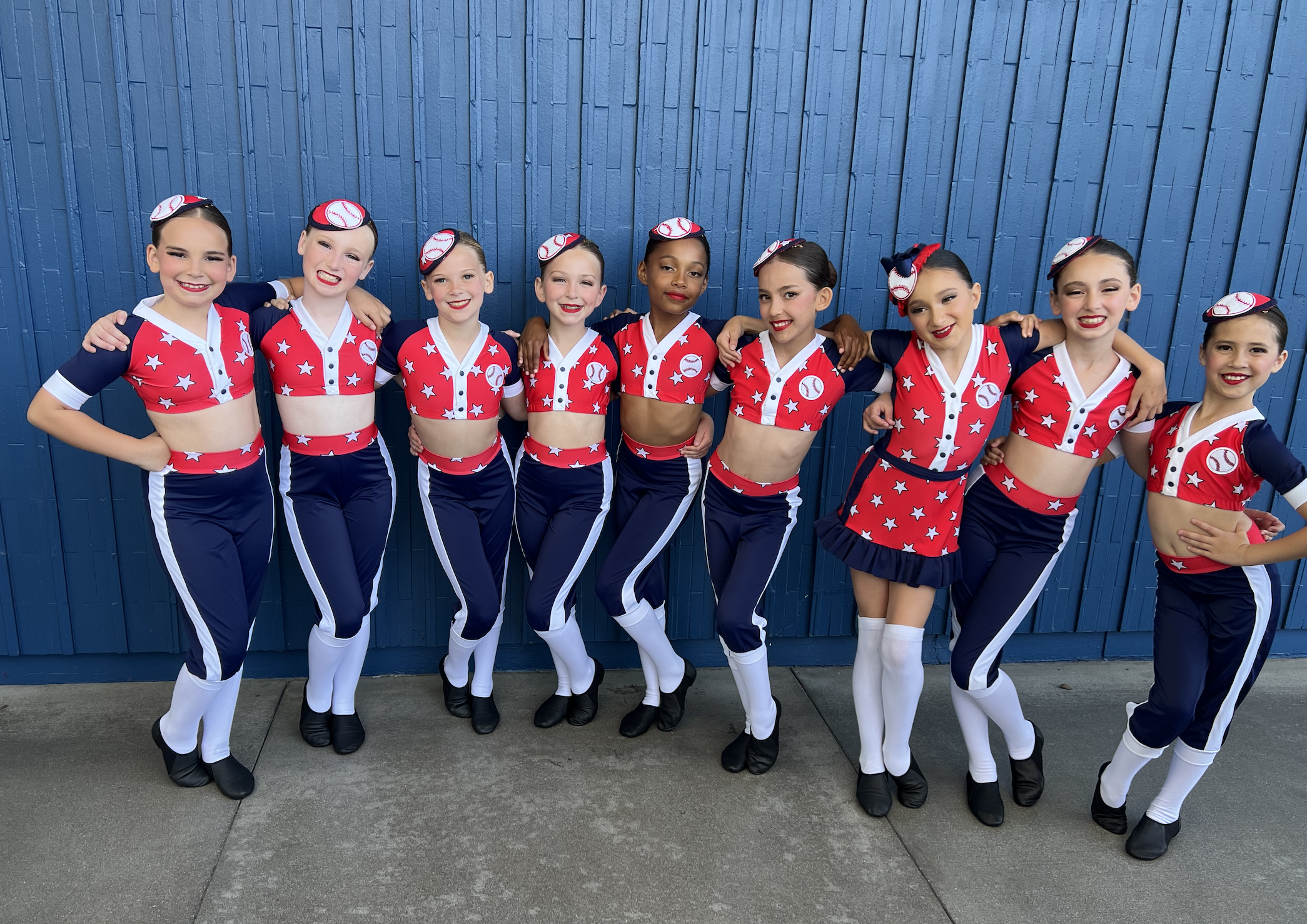 Group of nine young girls in cheerleader uniforms with baseball-themed hats, standing arm-in-arm against a blue wooden wall.