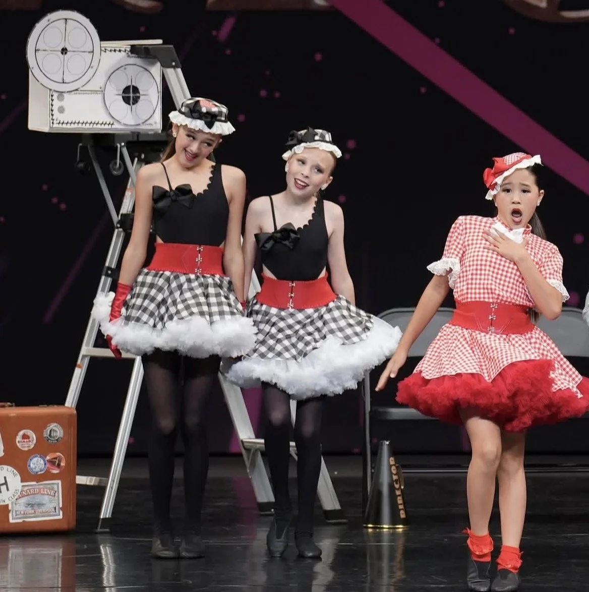 Three young girls performing on stage in vintage-style costumes. Two girls are dressed in black tops with checkered skirts, red belts, and headbands, while the third girl is dressed in a red and white checkered dress with matching headscarf. They app