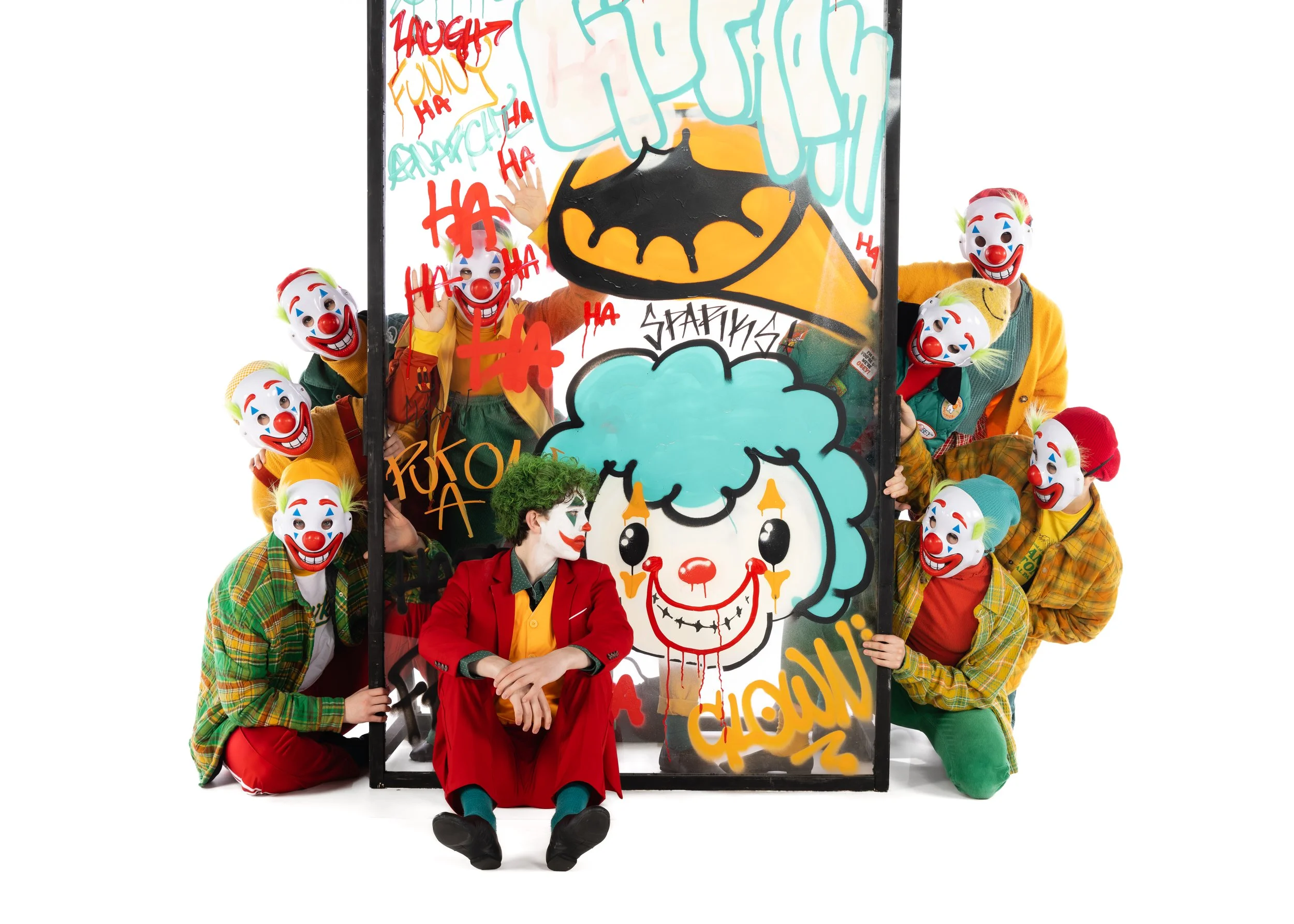 Group of people dressed as clowns posing around a large colorful artwork with graffiti and cartoonish drawings, including a smiling clown face and words like 'HA', 'ZACH', and 'FUN'.