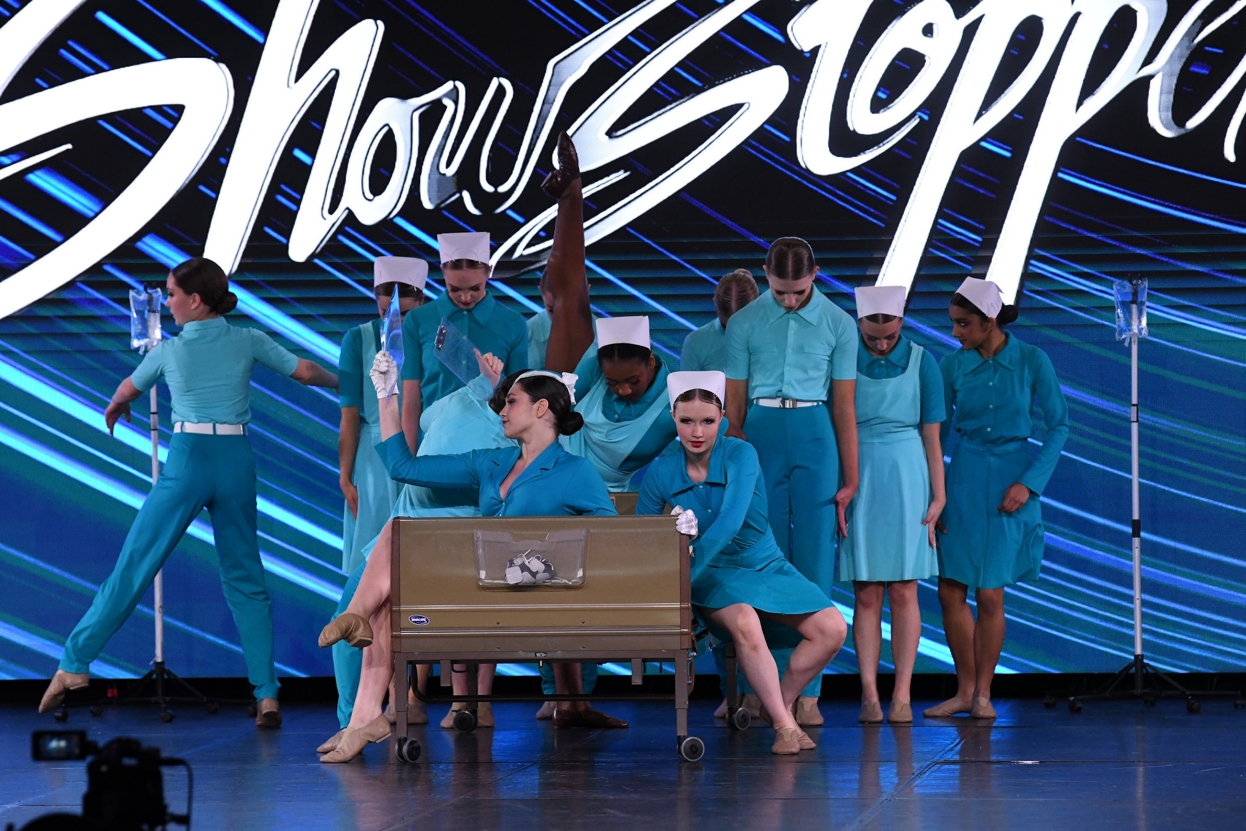 A group of performers dressed as nurses in teal uniforms and white caps, participating in a stage routine, with a large illuminated backdrop displaying the word 'Shonestock' in cursive, and a cardboard box with shoes in the foreground.