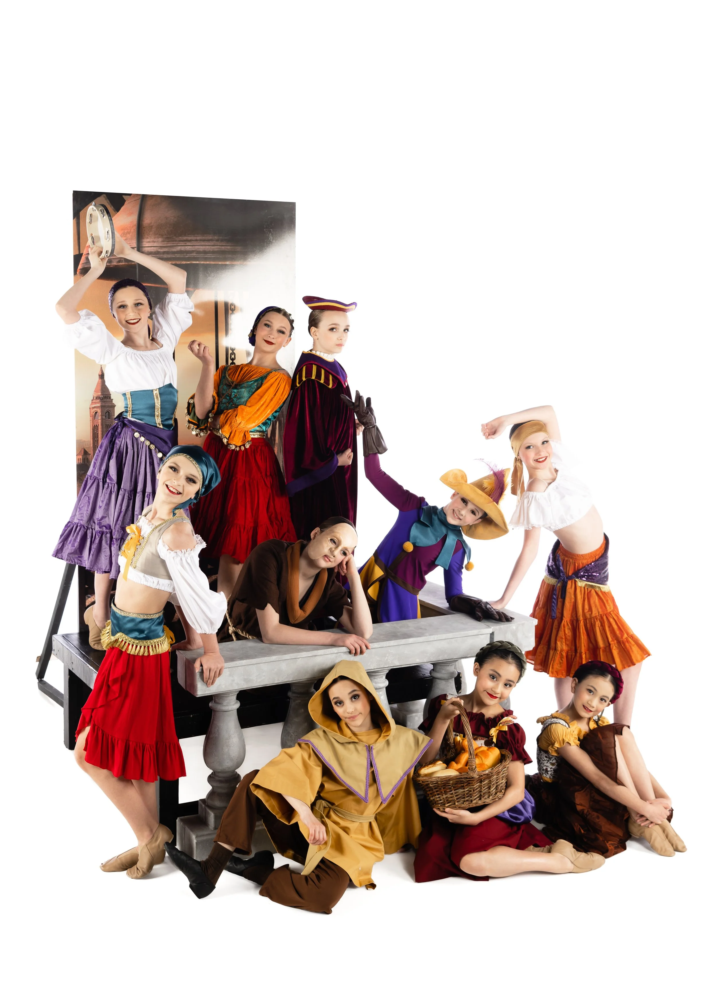 Group of children dressed in colorful costumes, posing around a gray stone-like structure, with some with props like a basket of bread. Background features a large poster or backdrop of Big Ben and London scenery.