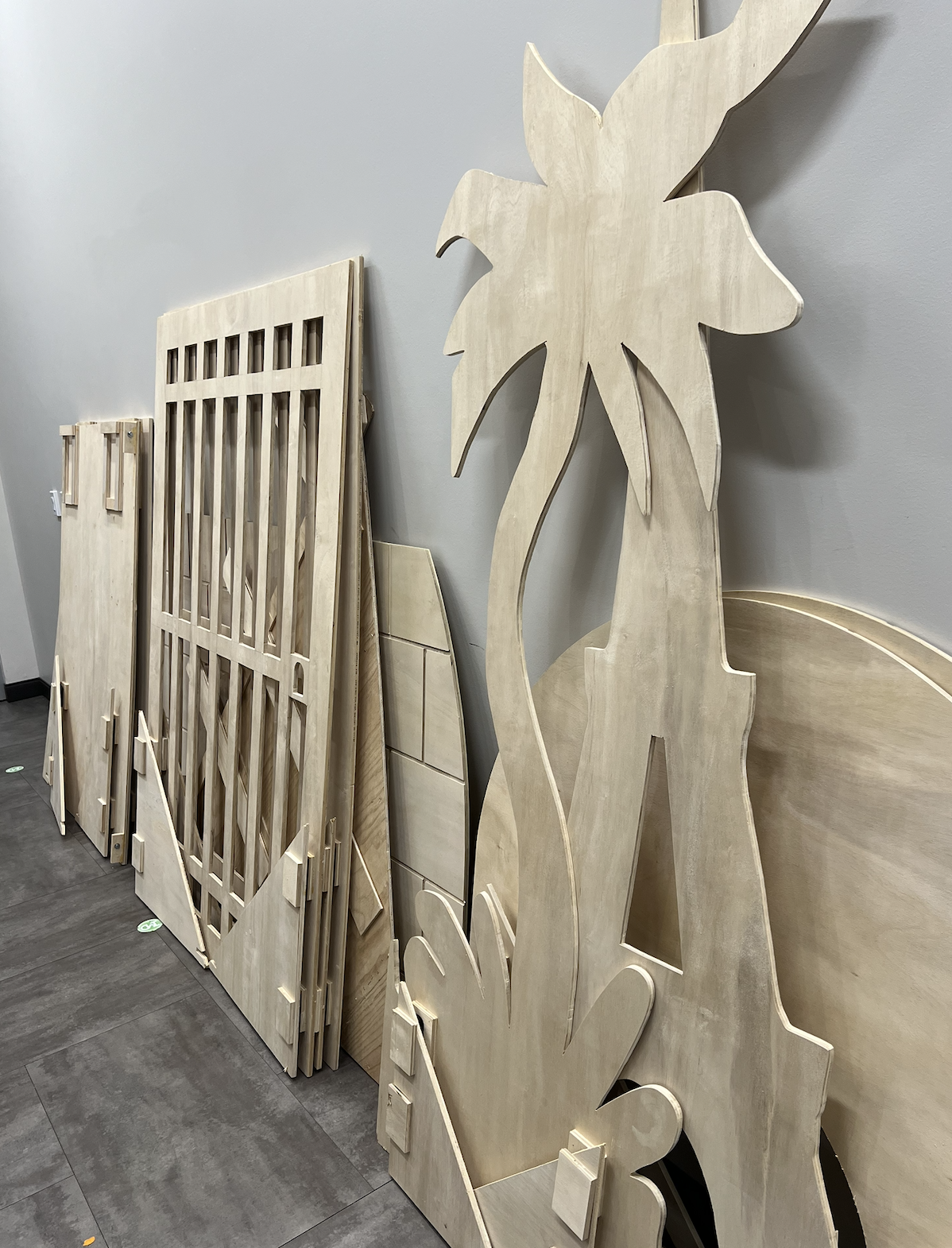 Stacked unfinished wooden decorative cutouts, including a large leaf shape, a picnic table, a small window frame, and a rectangular piece with holes, all leaning against a gray wall on a tiled floor.