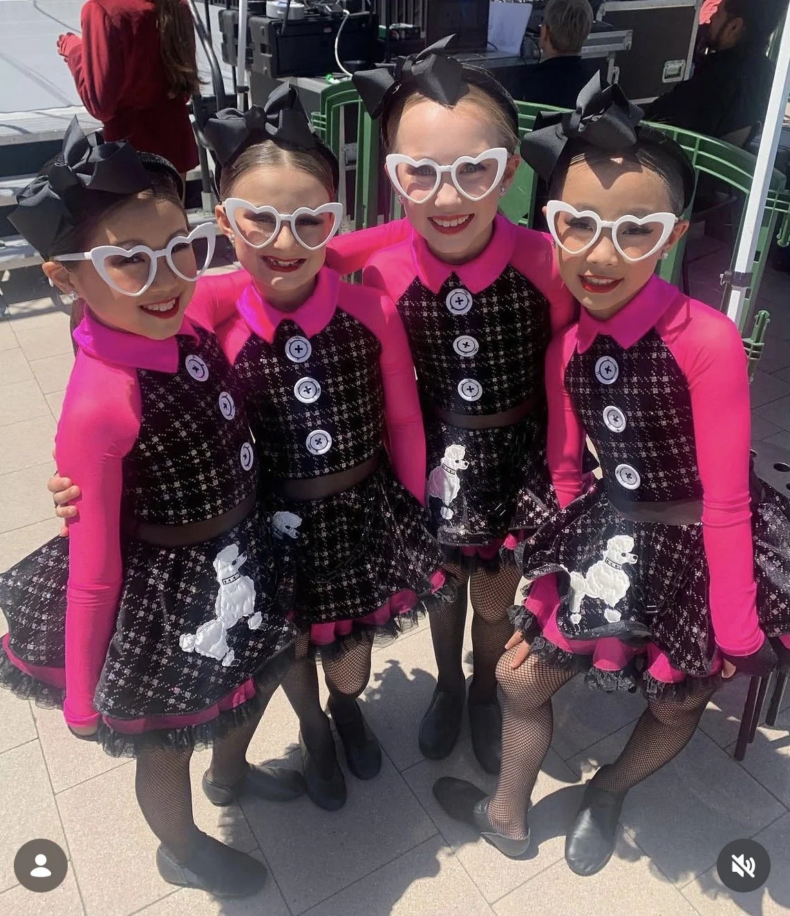 Four young girls standing close together, wearing matching black and pink dance costumes with poodle designs, black bow headbands, large white heart-shaped glasses, and fishnet tights, posing outdoors.