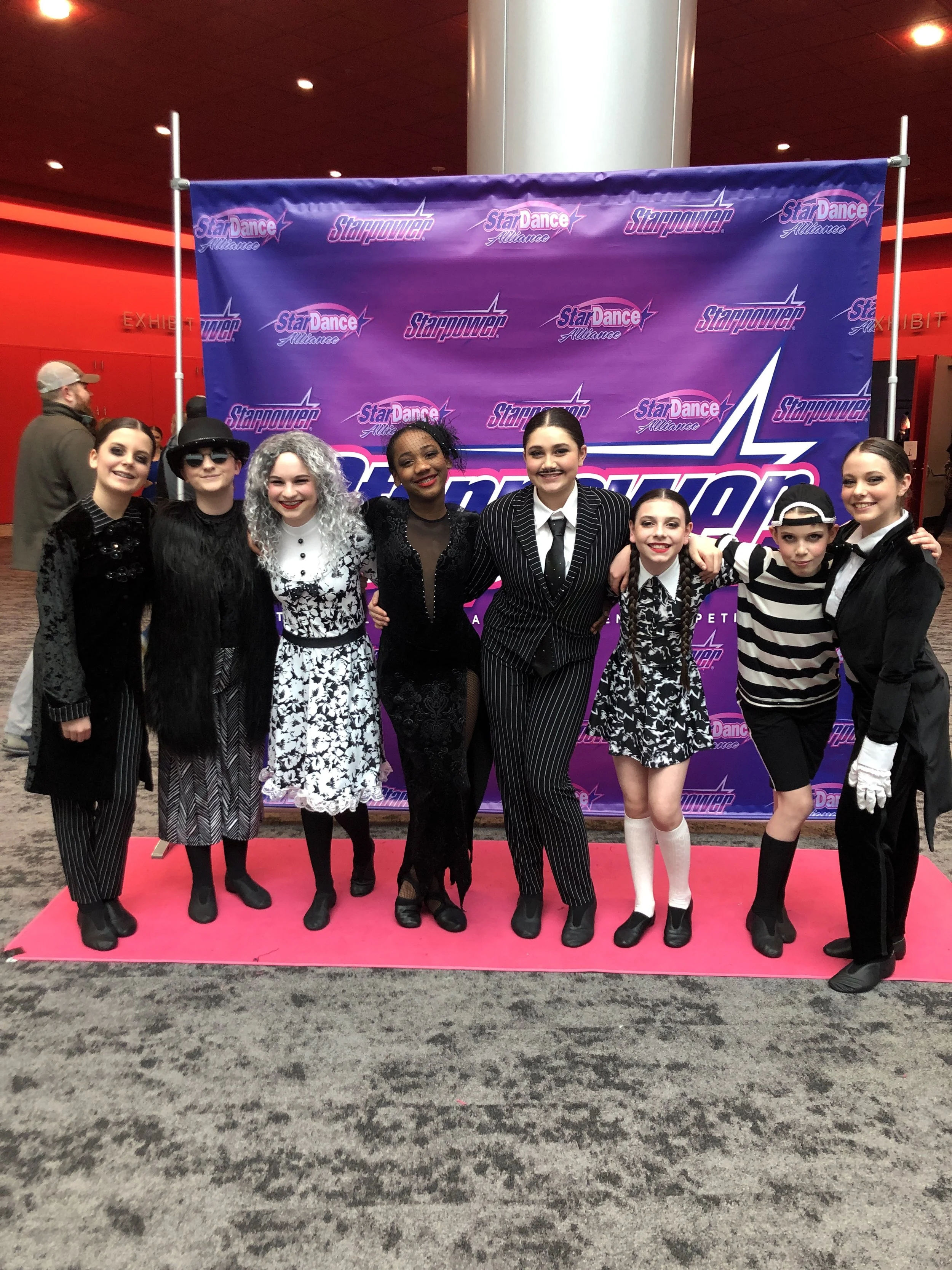 Group of eight young dancers in colorful costumes standing on a pink carpet in front of a purple backdrop with dance competition branding. They are smiling and posing together.