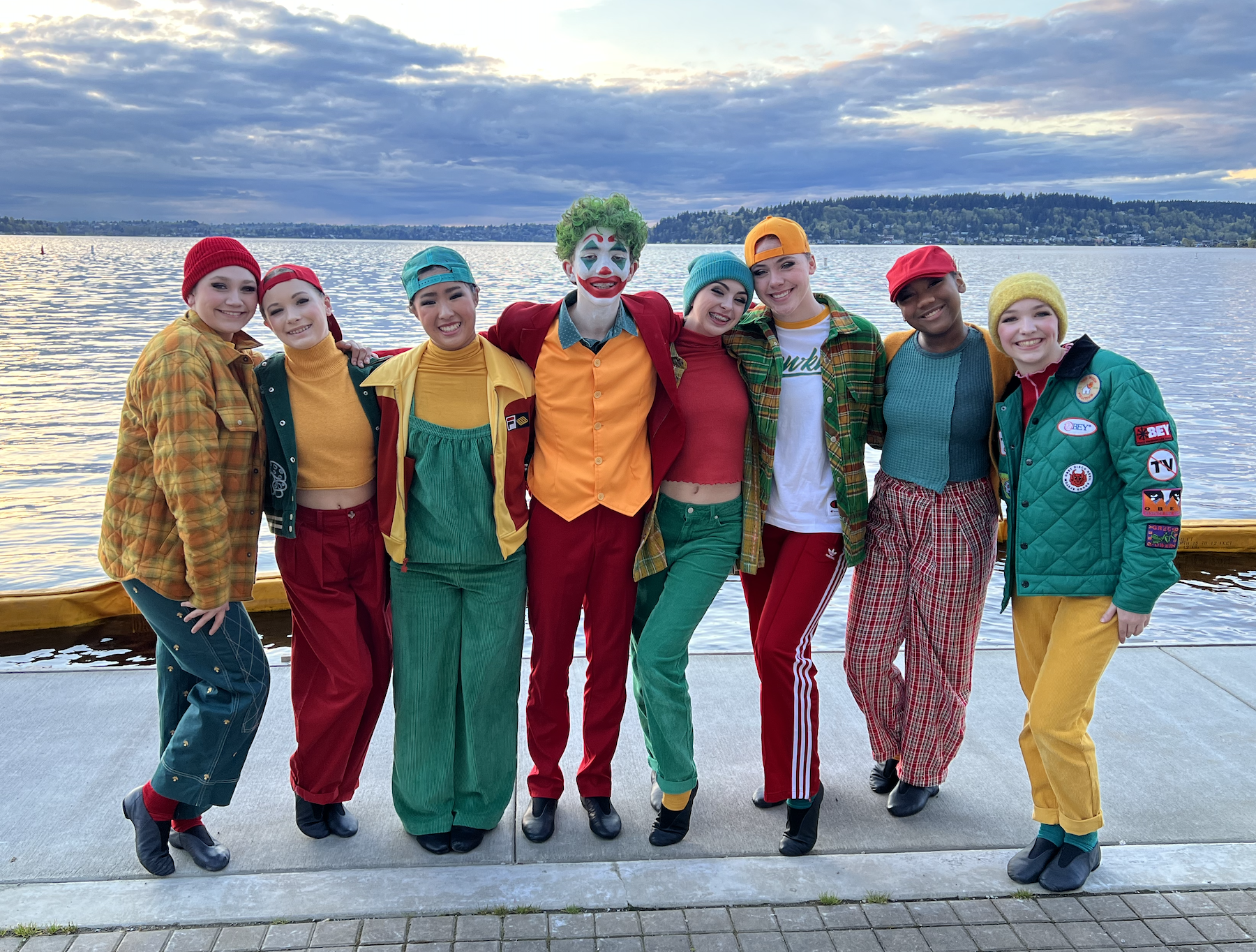 A group of nine people dressed as clowns, standing outdoors by a body of water with trees in the distance. They are smiling and have their arms around each other, wearing colorful outfits, some with hats and face paint.